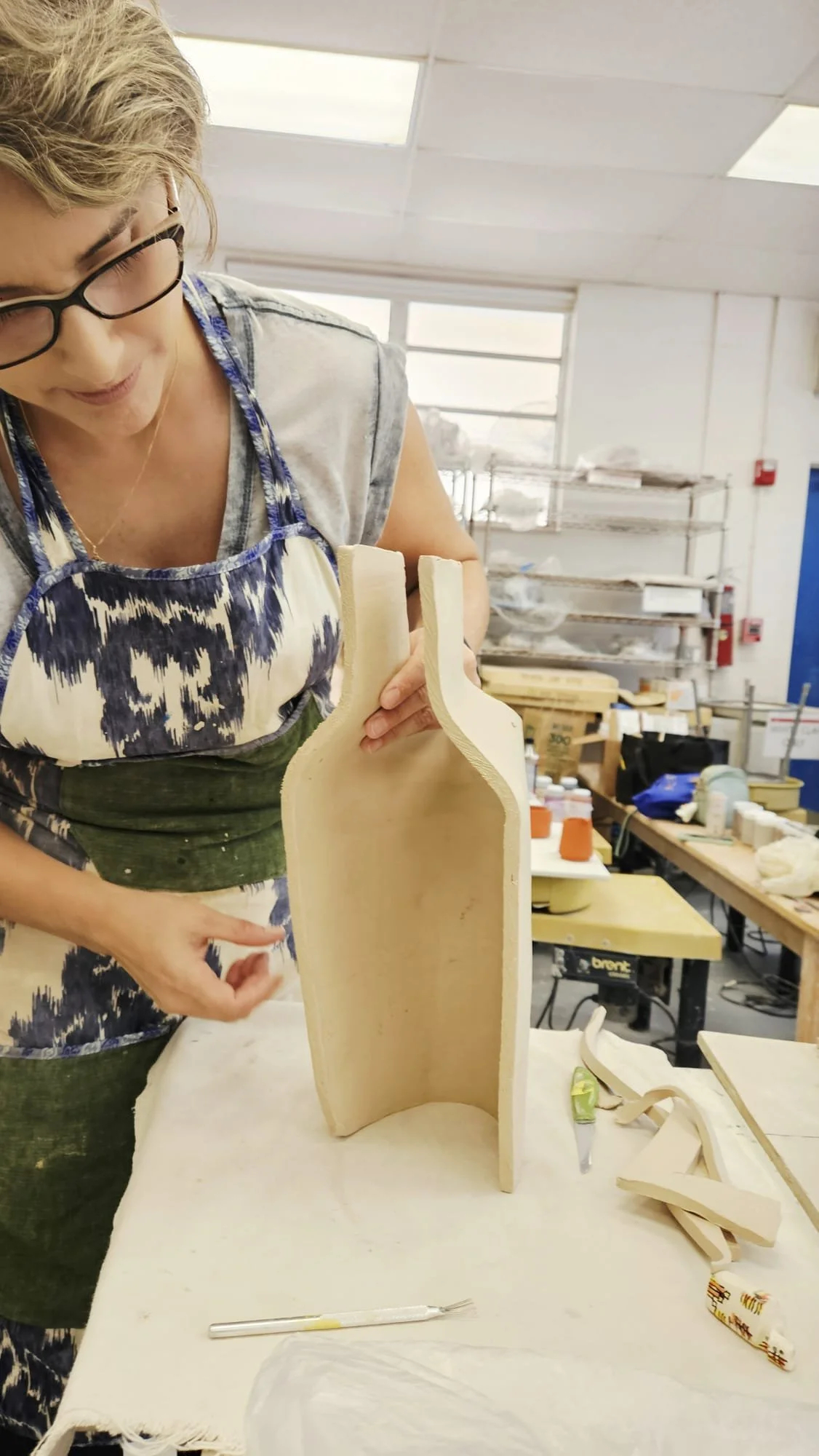 A woman working on a wooden sculpture in a workshop, surrounded by craft supplies, with shelves and windows in the background.