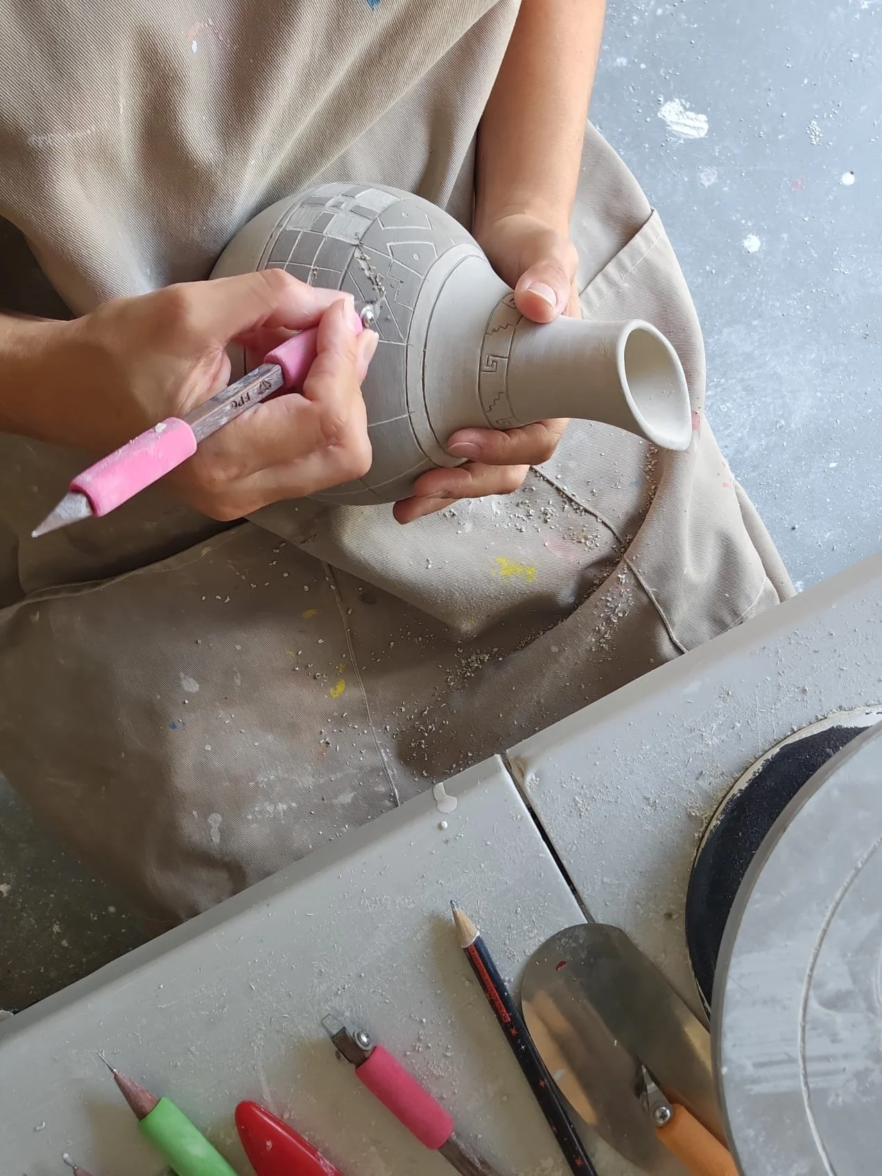 Person carving a pre-hispanic design into a ceramic vase with a pink carving tool, wearing an apron and having carving and pottery tools nearby.