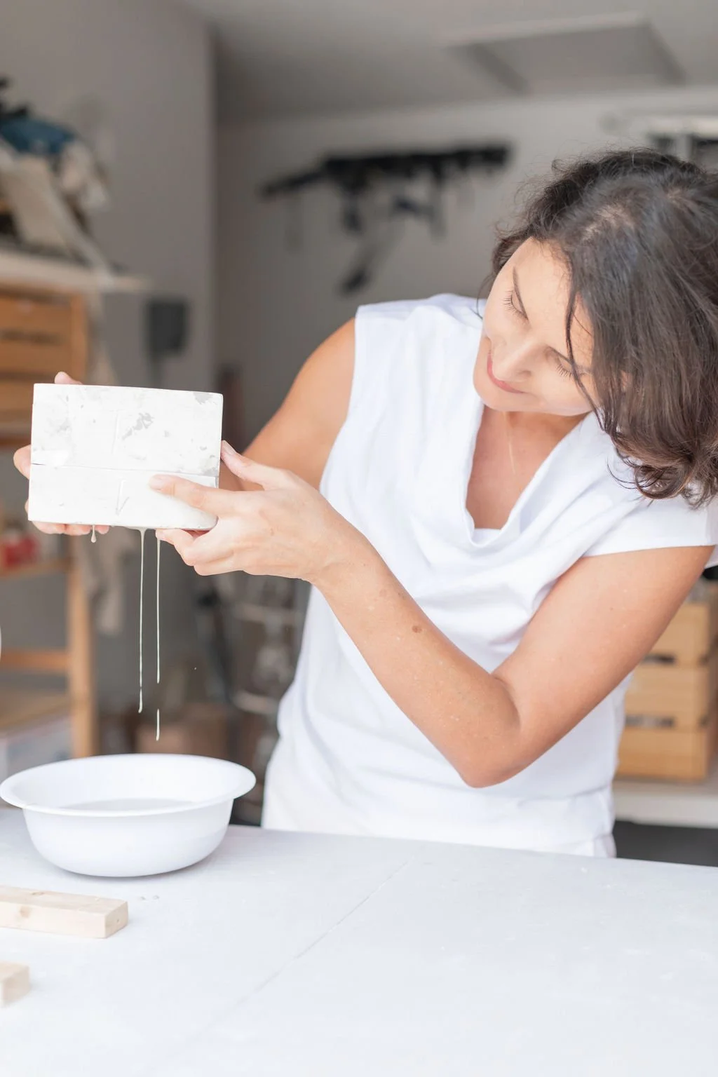 Woman holding a dripping block of cheese over a white bowl in a kitchen or workshop.