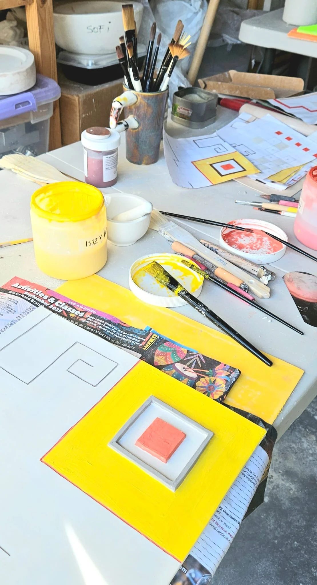 Art supplies on a table, including paintbrushes in a container, paint jars, paper with geometric designs and colorful paintings, and tools for painting.