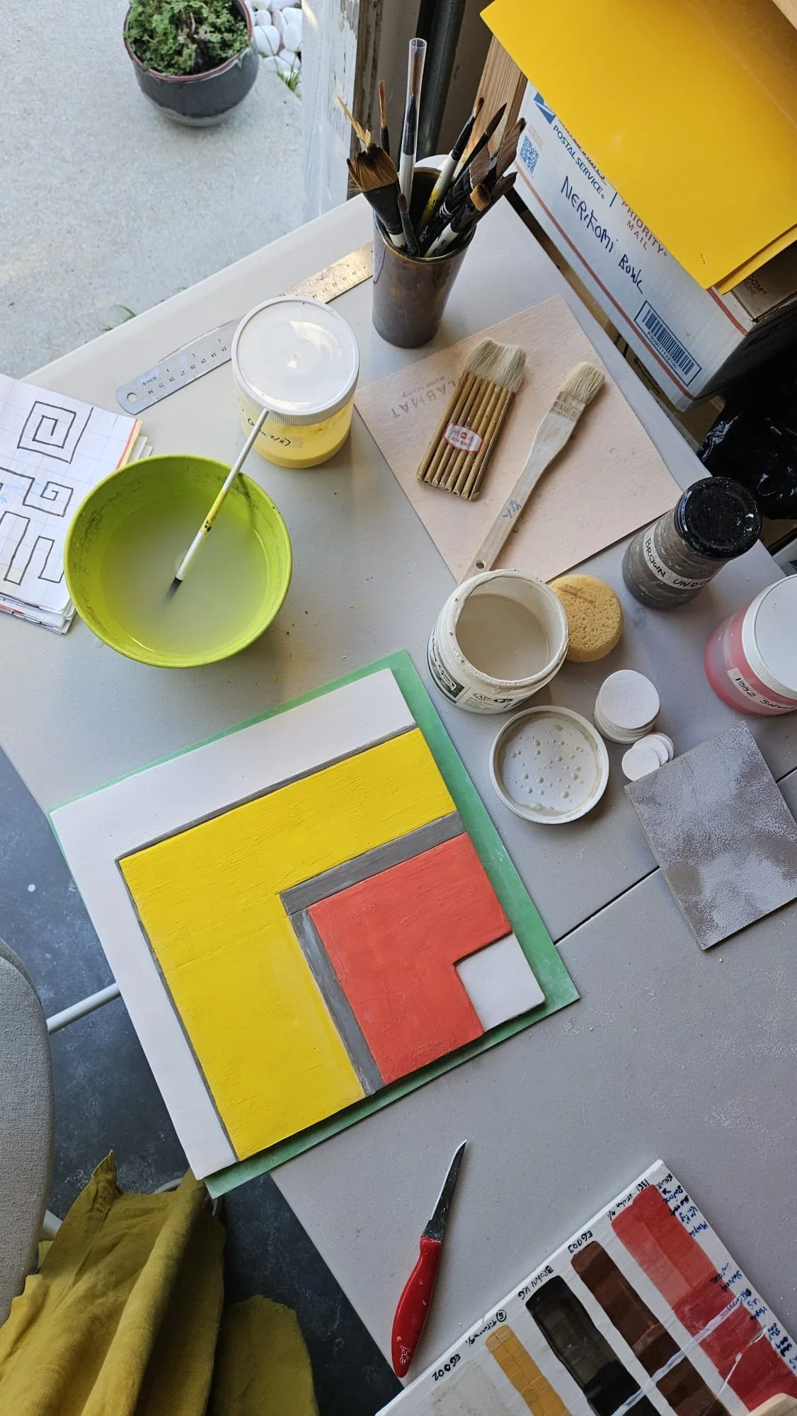 A workspace with art supplies including paints, brushes, a color palette, and a painting of concentric squares in yellow, red, black, and gray on a white surface. There are also paper, a ruler, and a container with water or a liquid for cleaning brus