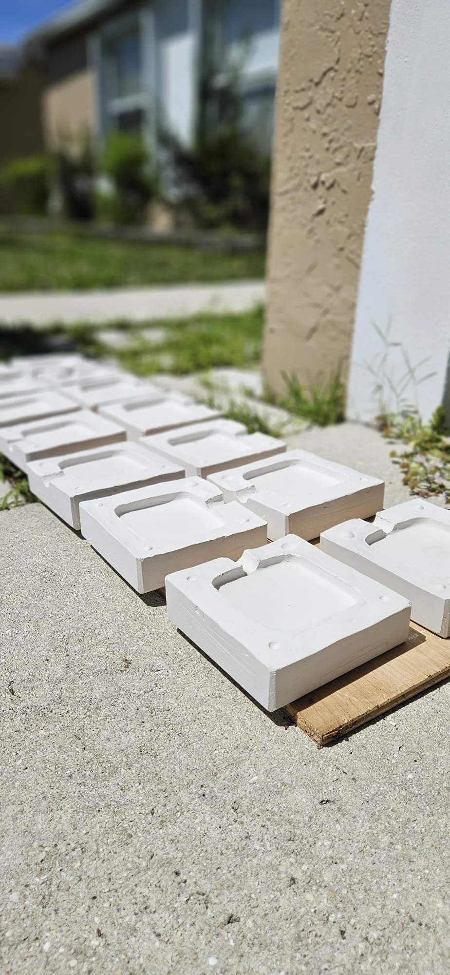 Several white rectangular ceramic tiles with handmade textures are laid out on a concrete surface outside, next to a beige and white wall with some grass nearby.
