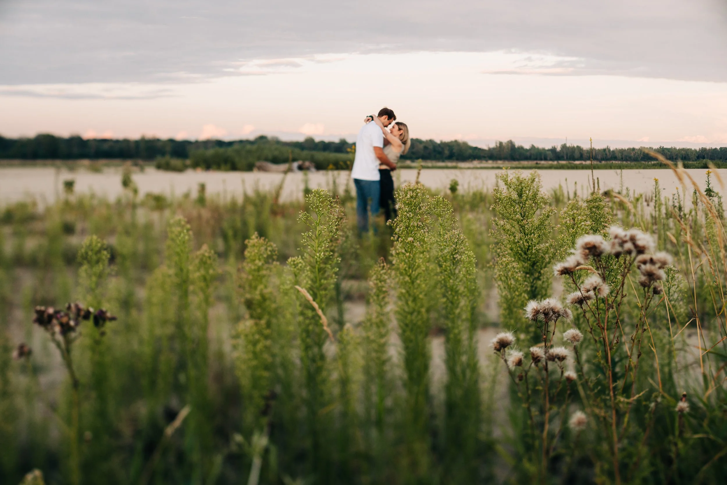 A couple embraces in a field with green plants, with a lake and cloudy sky in the background.