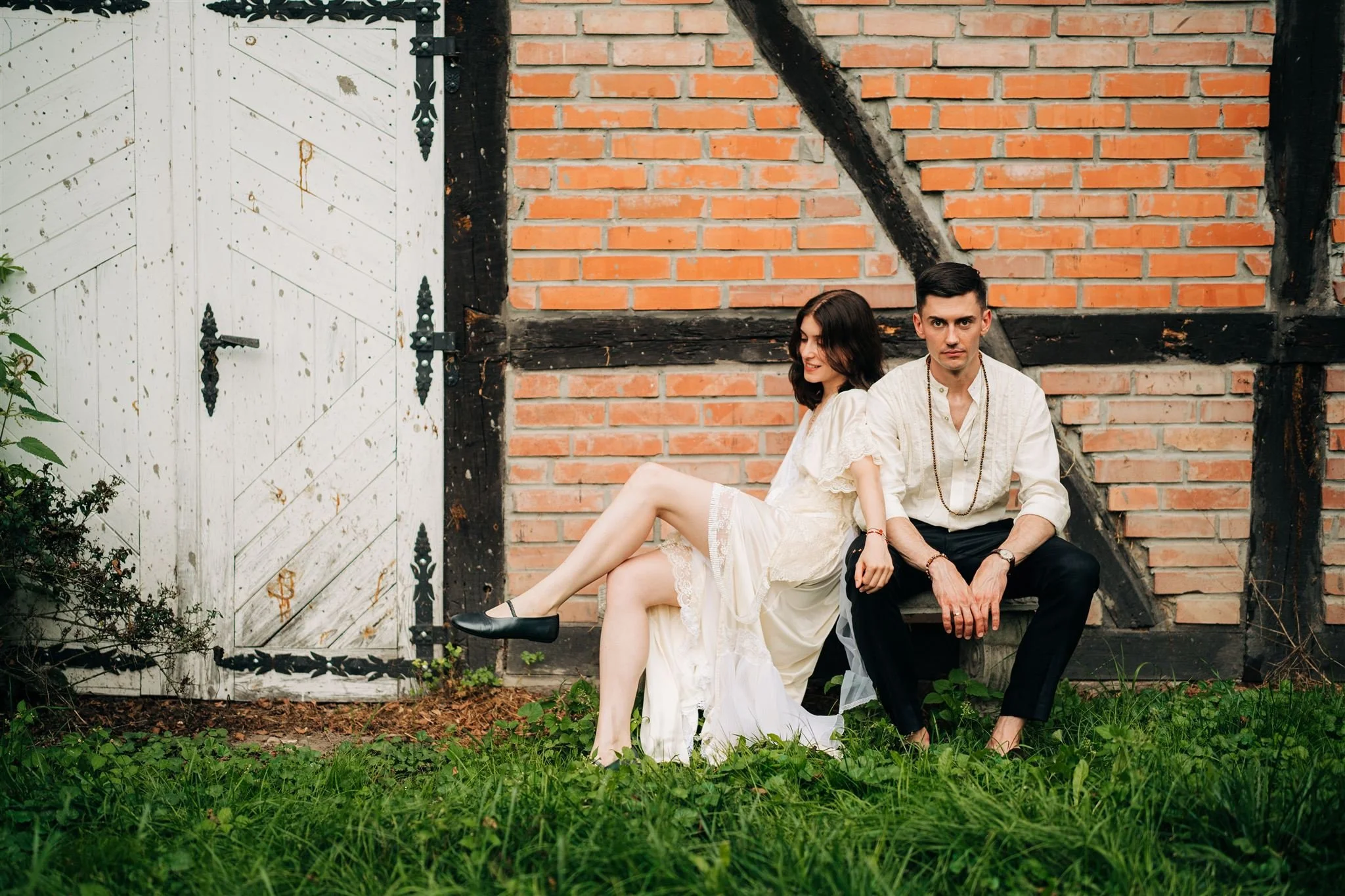 A young couple sitting outdoors against a rustic brick wall with a white wooden door, with green grass in the foreground. The woman wears a white dress and black shoes, and the man wears a white shirt and black pants.