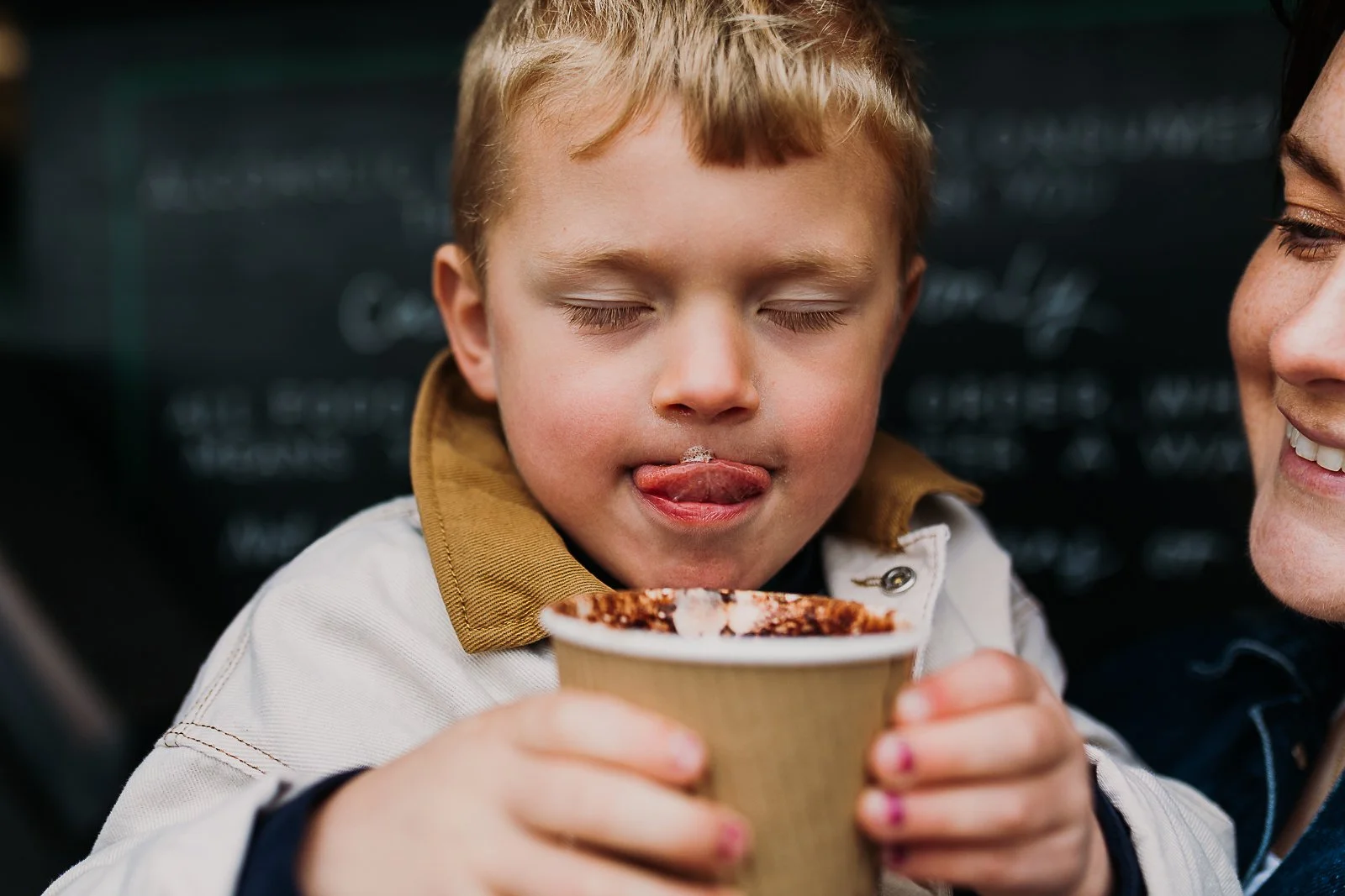 Young boy drinking hot chocolate while his mum smiles at him, while having a cuddle with his mum in Whitstable.