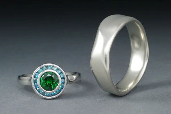 Engagement ring in white gold with a bezel set green tsavorite garnet surrounded with a channel set halo of blue diamonds.  Men's band in white gold in an organic sculptural form.