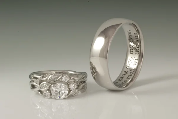 White gold set. Her rings feature accent diamonds set in floral inspired forms twisting around and flanking the center stone.  His ring features inside engraving of a personalized quote. 