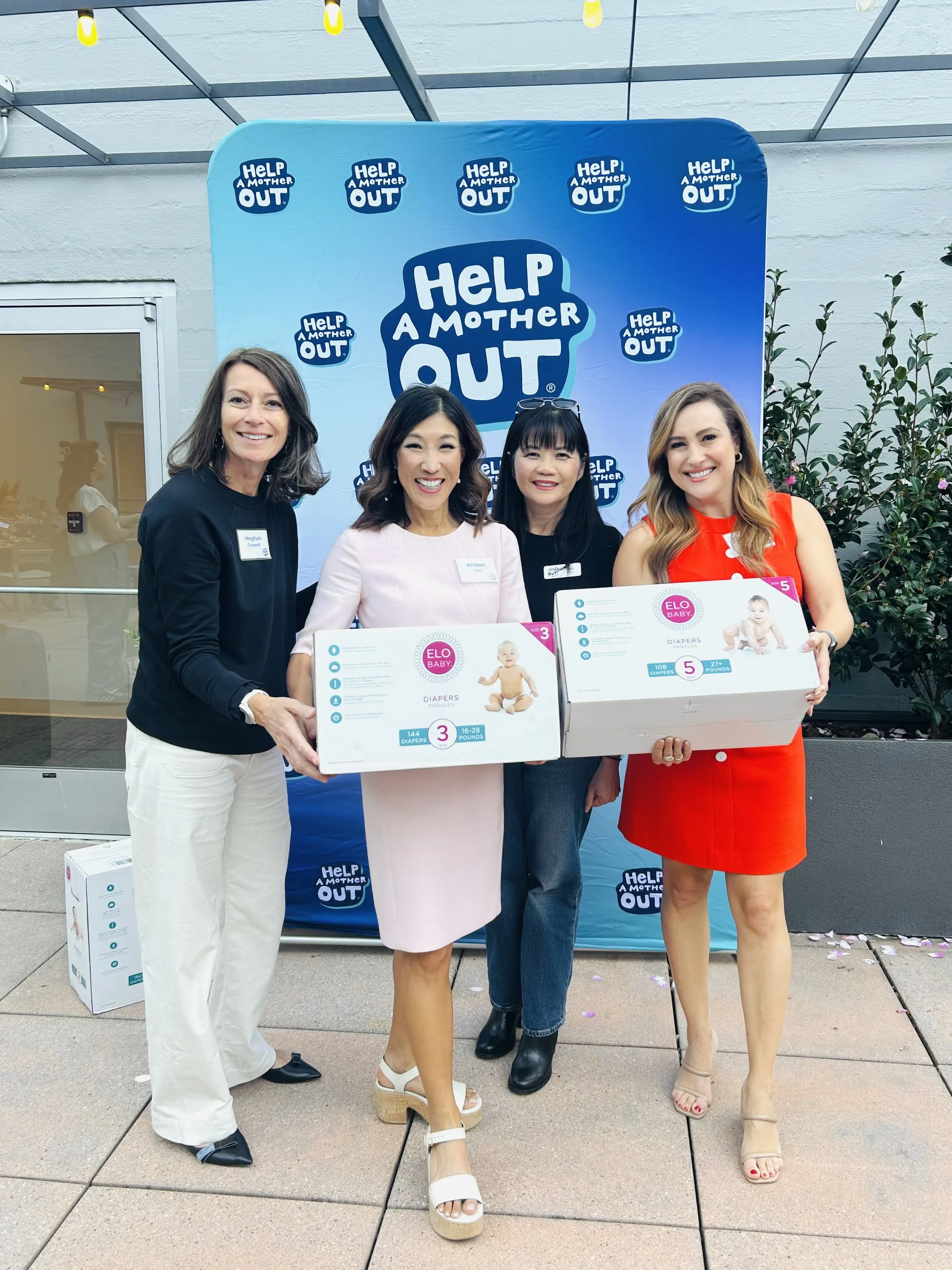 Meghan Crowell, Kristen Sze, Lisa Truong, and Karina Nova pose in front of a Help a Mother Out backdrop on a rooftop patio. Kristen and Karina are holding up boxes of ELO Baby diapers.