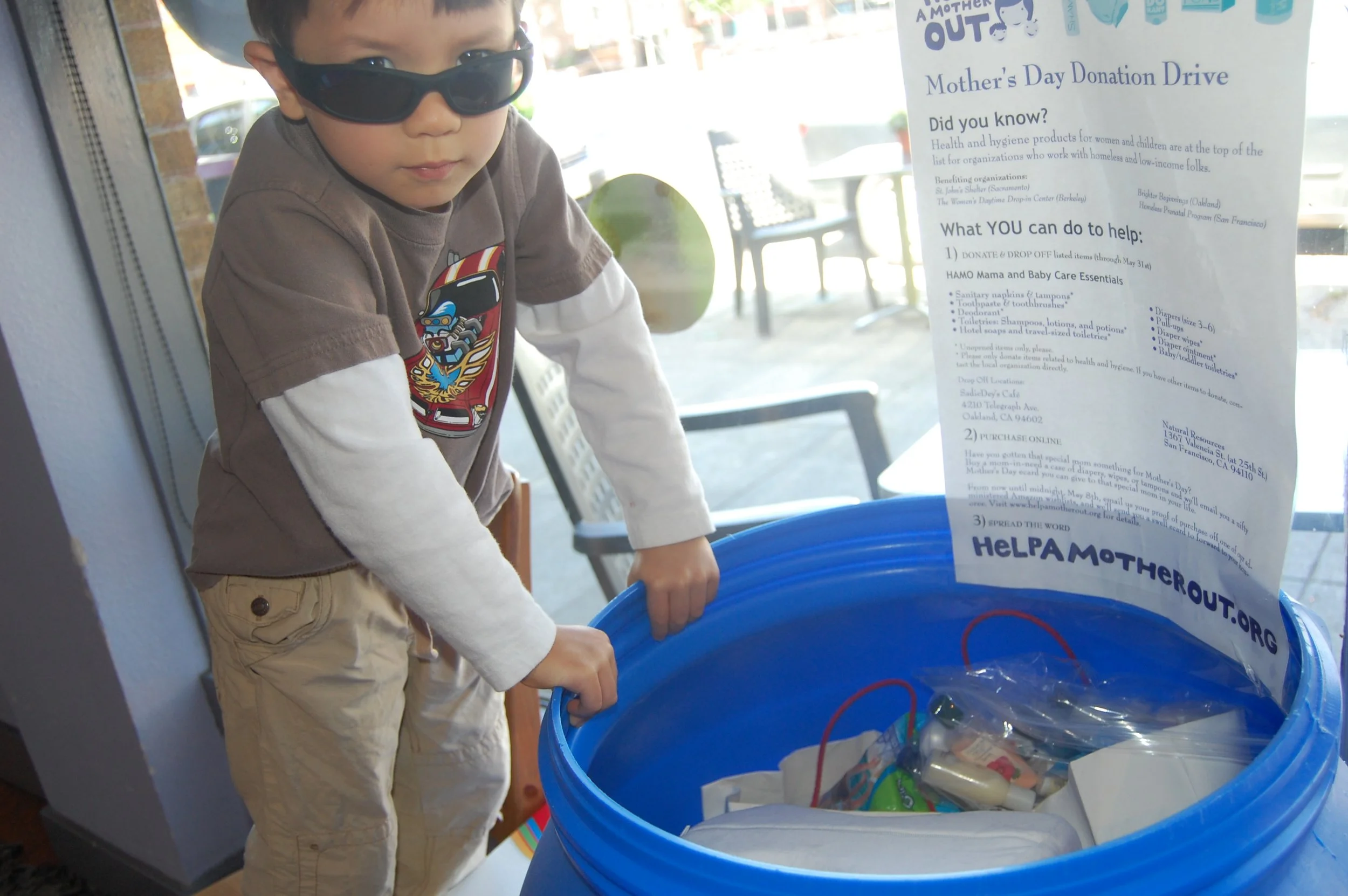 A young child stands beside a blue donation bin filled with care items at a Help a Mother Out Mother's Day donation drive collection site.
