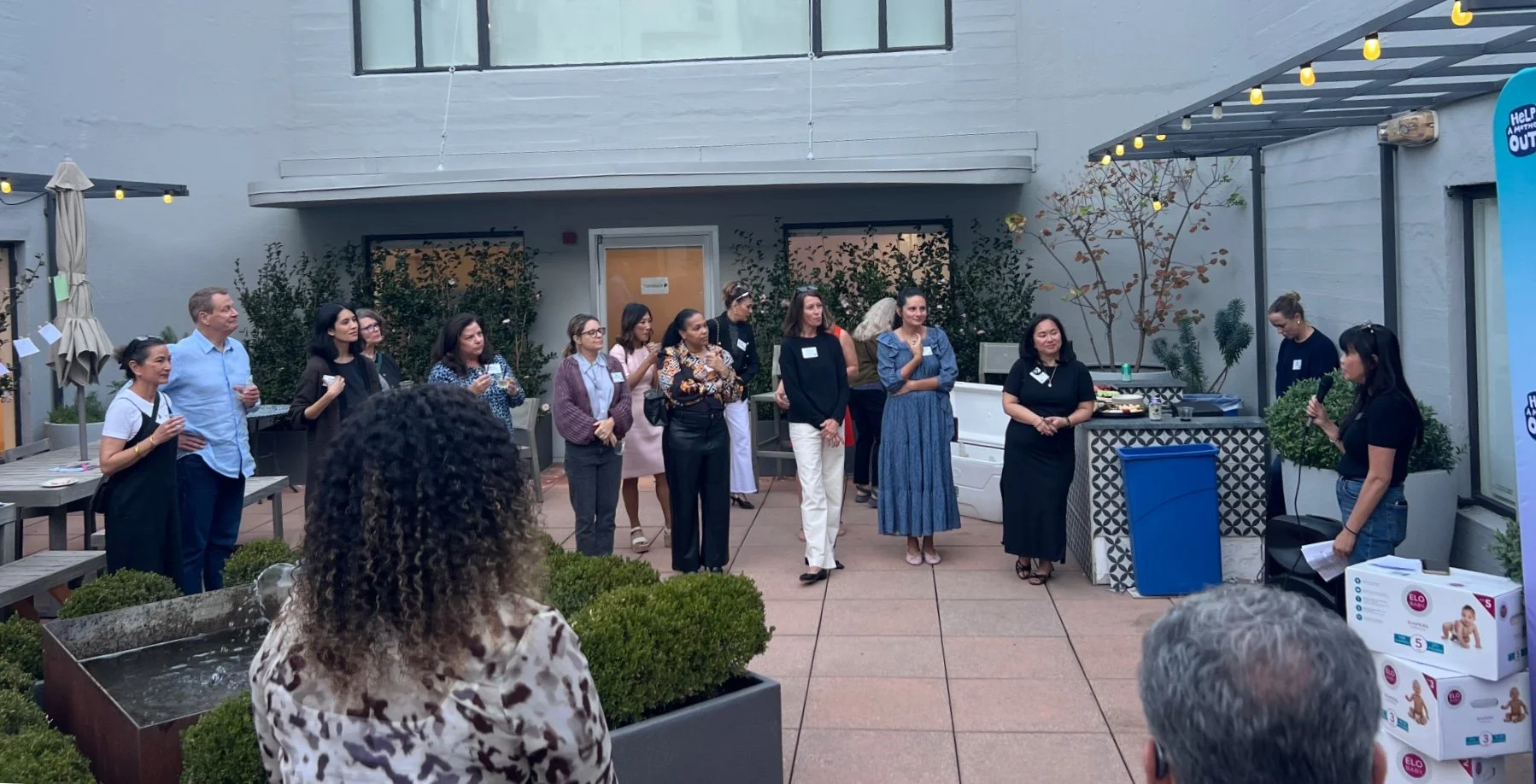 Lisa Truong holds a microphone and notes and talks to a semicircle of guests with name tags on a rooftop patio. String lights hang overhead, cards are clipped along a line at the left, and boxes of ELO Baby diapers are stacked in the foreground.