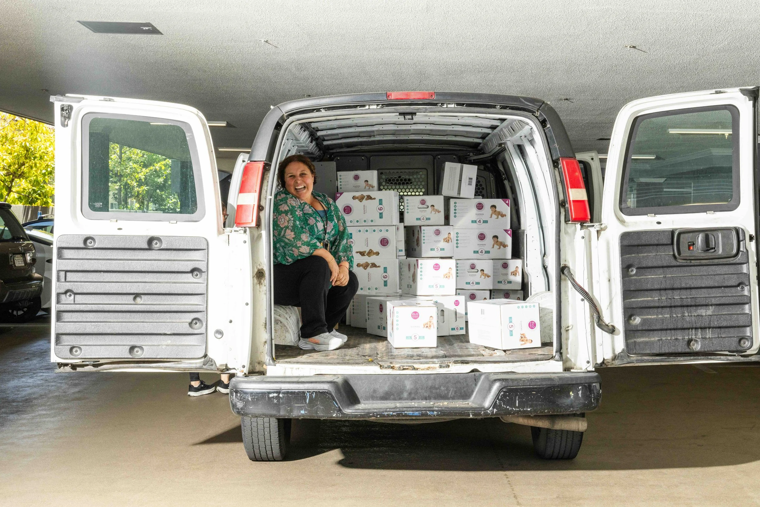 A woman sits in the back of a cargo van loaded with stacked boxes of diapers staged for community distribution.