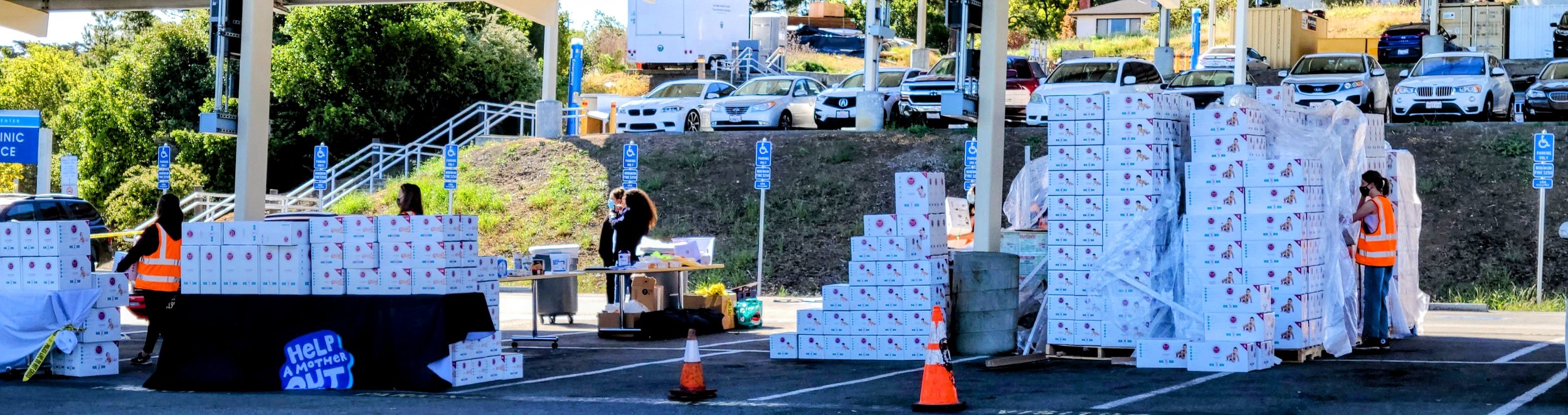Help a Mother Out staff in safety vests run a large diaper distribution at a clinic site, with pallets of boxed diapers staged across the parking lot.