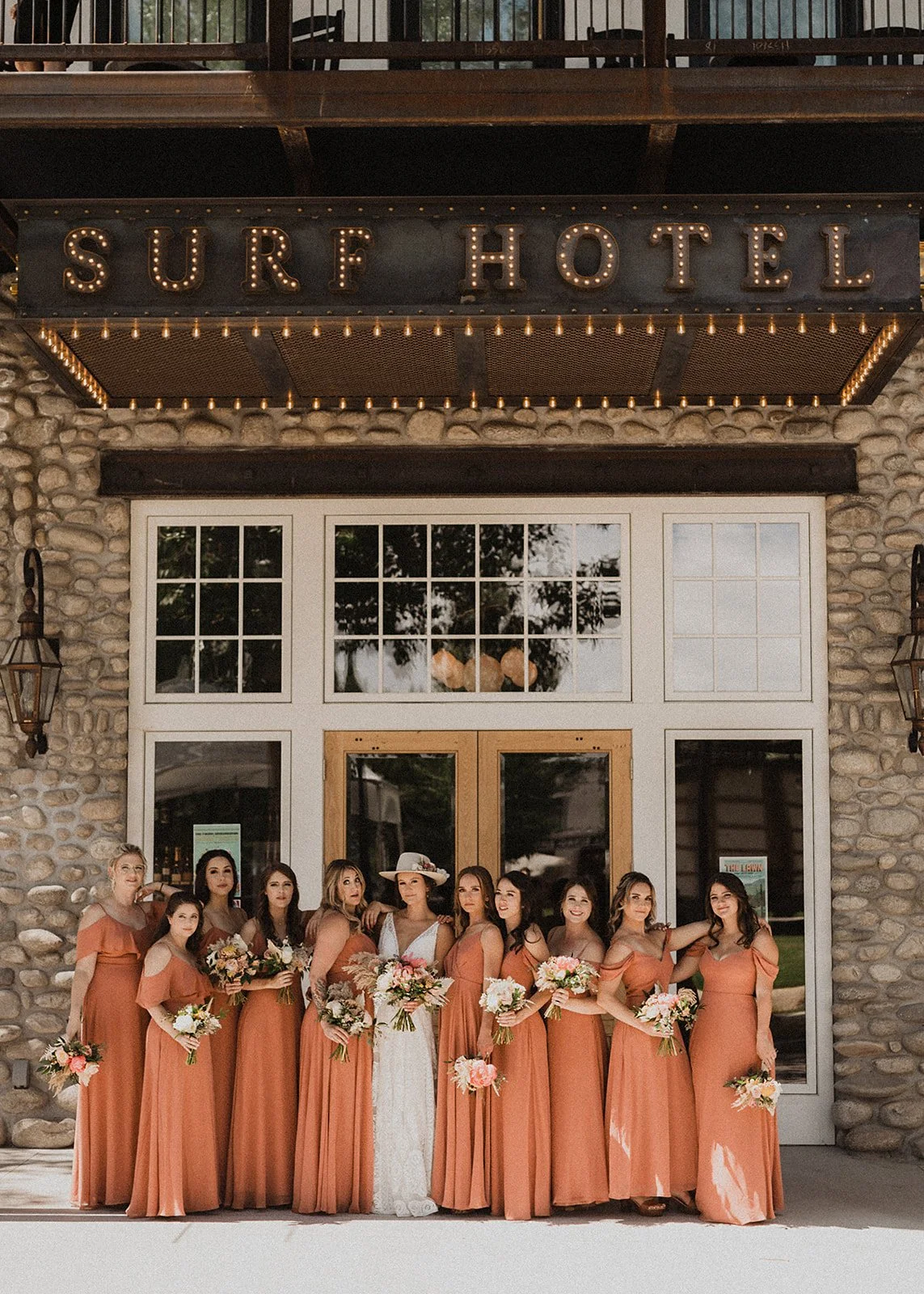 Group of bridesmaids in coral dresses and a bride in a white gown with flowers in front of the Surf Hotel entrance.