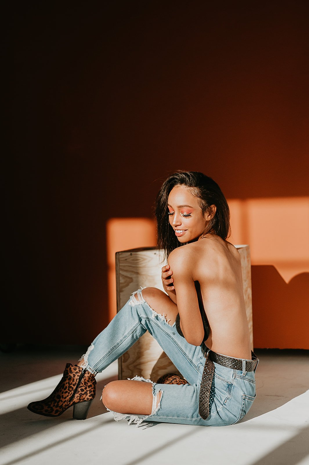 A person sitting on the floor wearing ripped jeans and leopard print boots, with a wooden box in the background, against a brown wall.