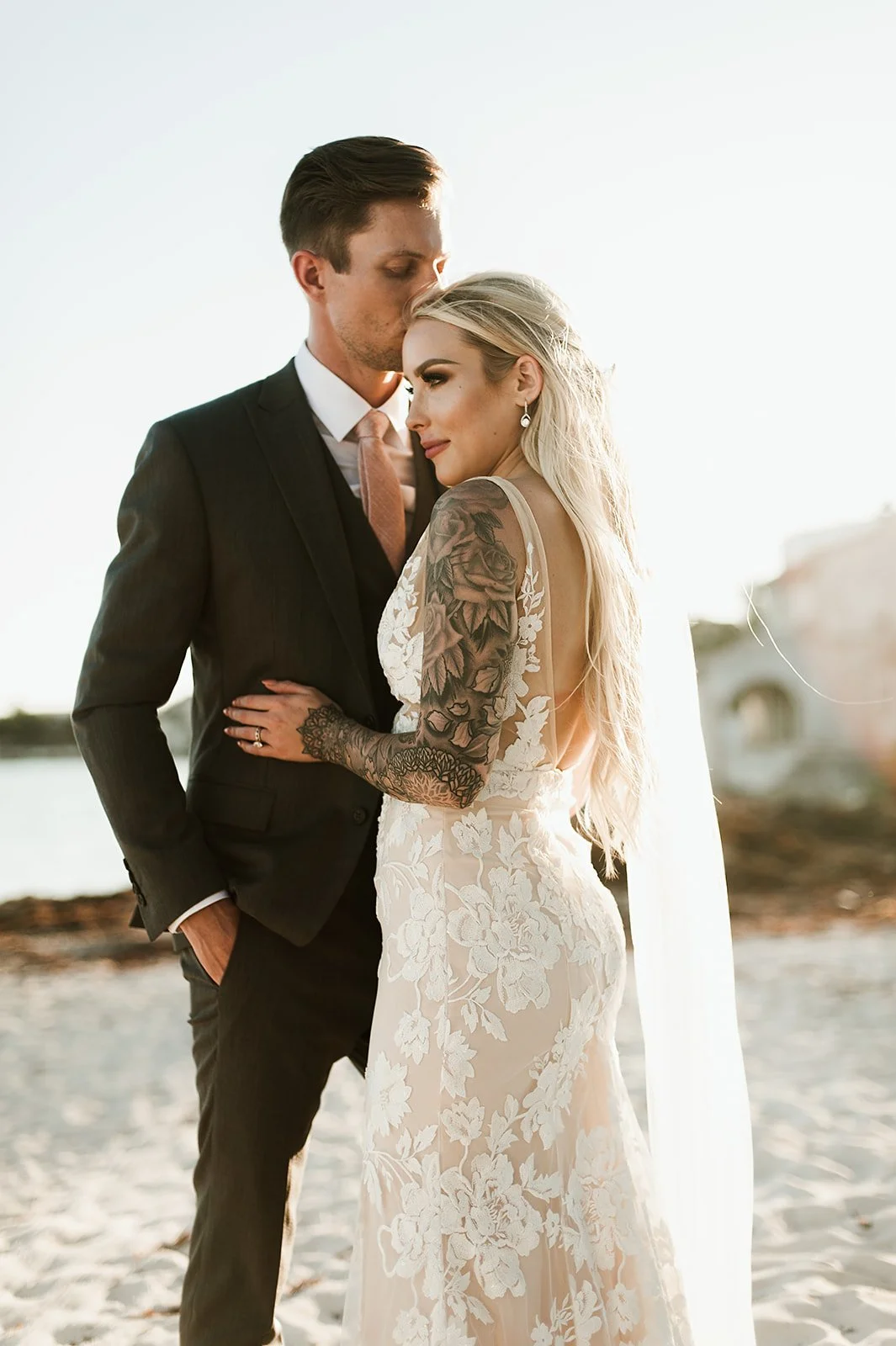 Bride in lace gown with floral tattoo, standing with groom in suit on beach.