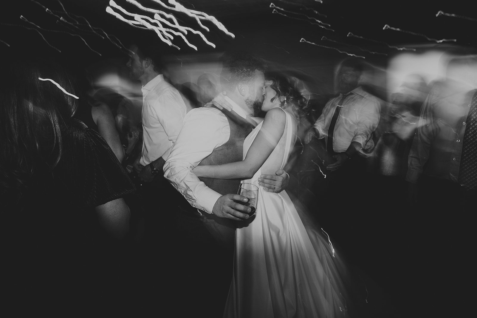 Black and white photo of a couple dancing closely at a party, with blurred figures and light streaks in the background.