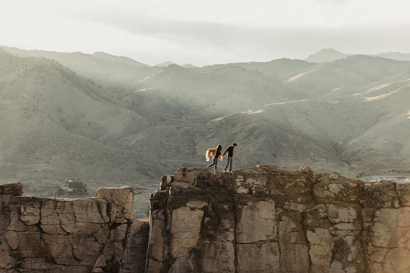Two people walking on rocky cliff edge with misty mountains in the background.