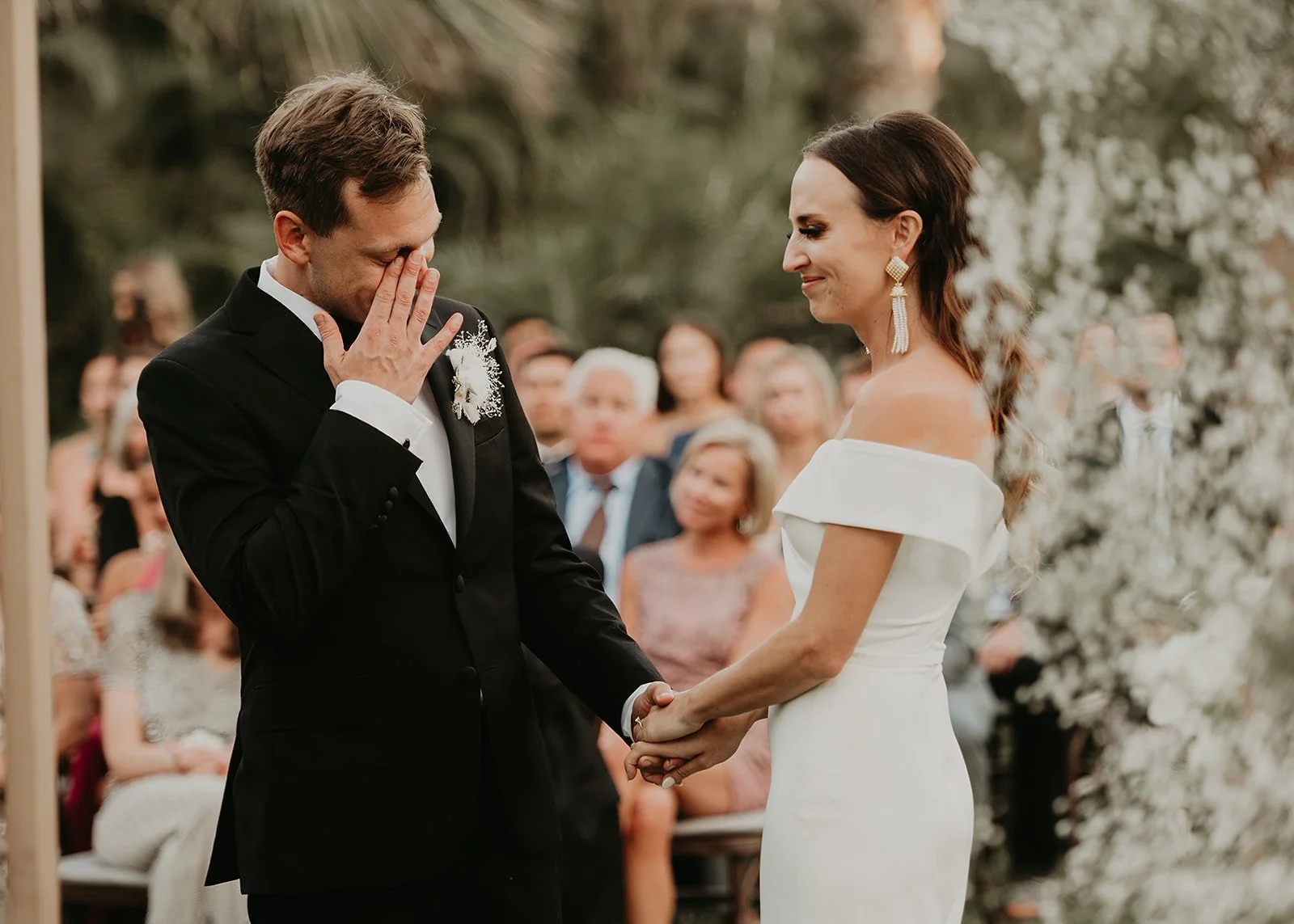 A man and woman standing and holding hands during an outdoor wedding ceremony. The man is wearing a black suit with a white shirt, raising a hand to his face. The woman is wearing a white off-shoulder dress, smiling gently. In the background, guests 