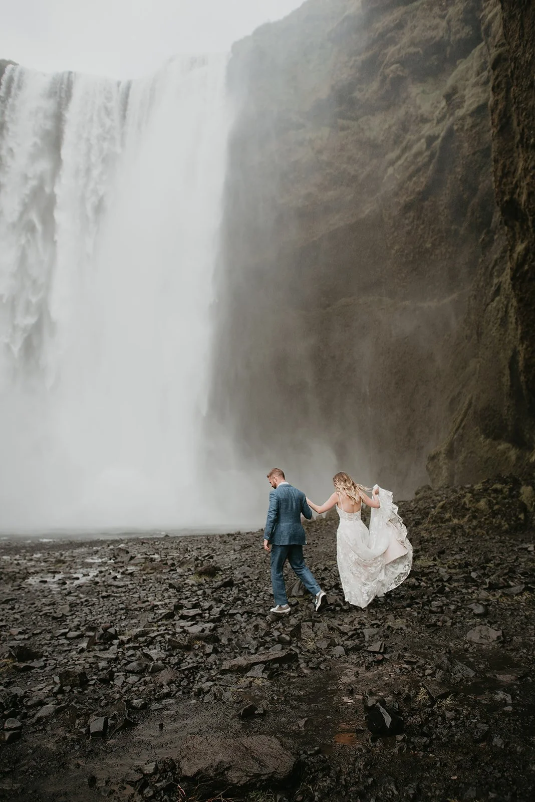 A couple in wedding attire walking near a large waterfall on a rocky terrain.