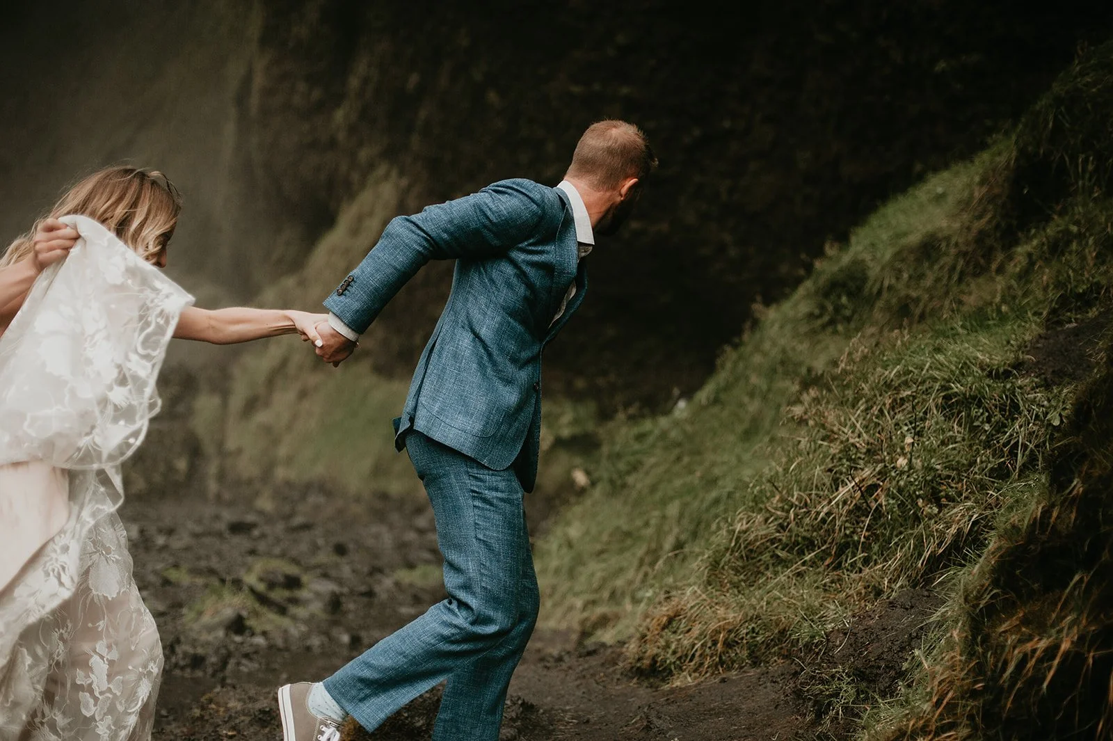 A couple in formal attire walking outdoors, with the man leading the woman, who is holding a lace veil.
