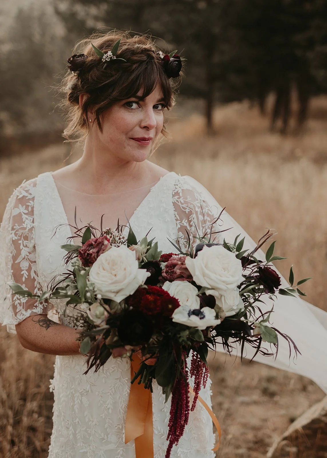 Bride holding bouquet with roses and greenery, wearing lace dress, outdoor setting.