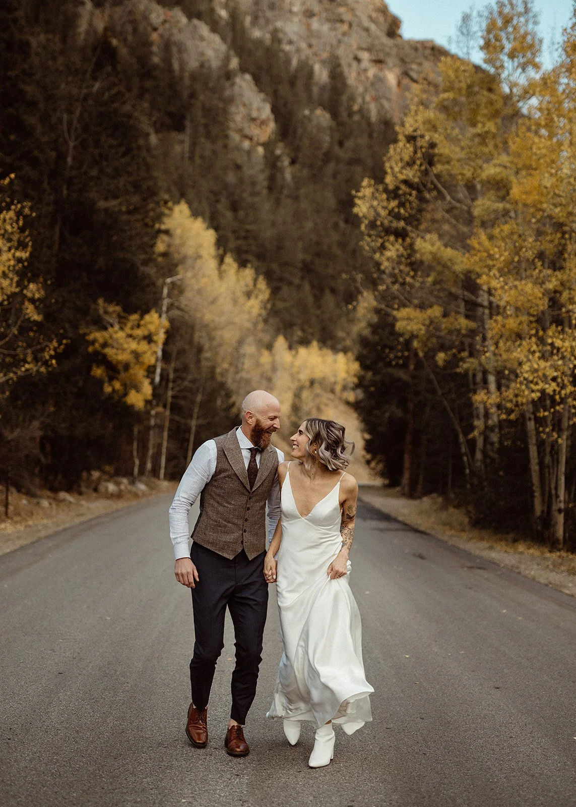 A couple in wedding attire smiling and walking down a tree-lined road in an autumn setting.