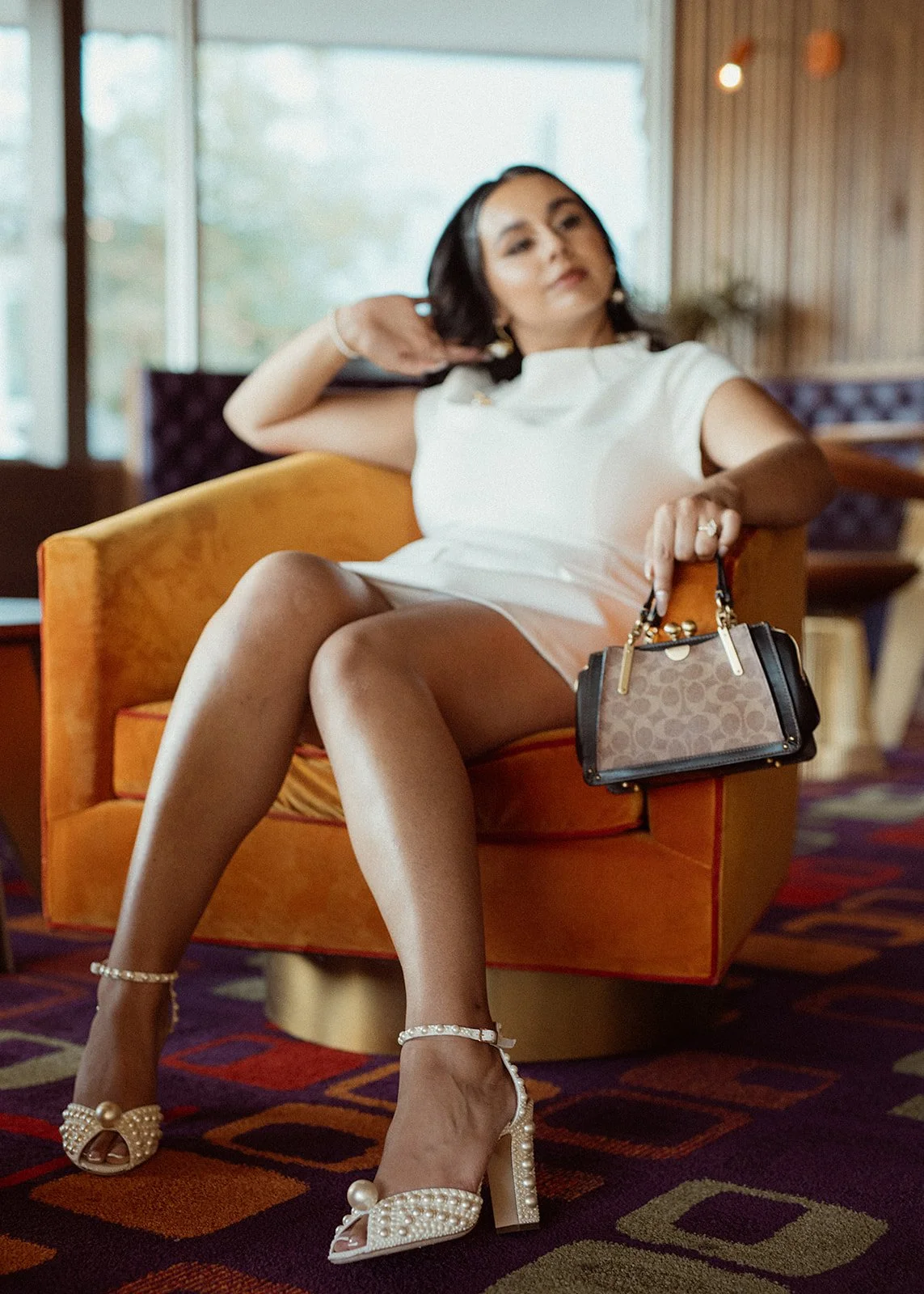Woman in white dress and pearl heels sitting on orange chair, holding handbag.