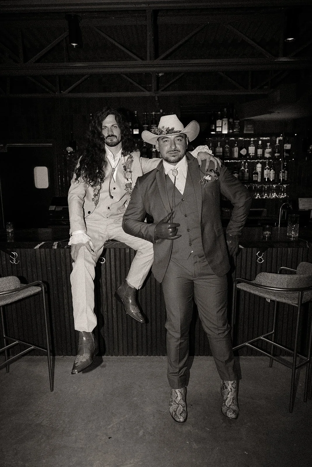 Two men in formal outfits, one with long hair sitting on a bar counter, the other wearing a cowboy hat and standing, in a bar setting with bottles in the background. Image is in black and white.