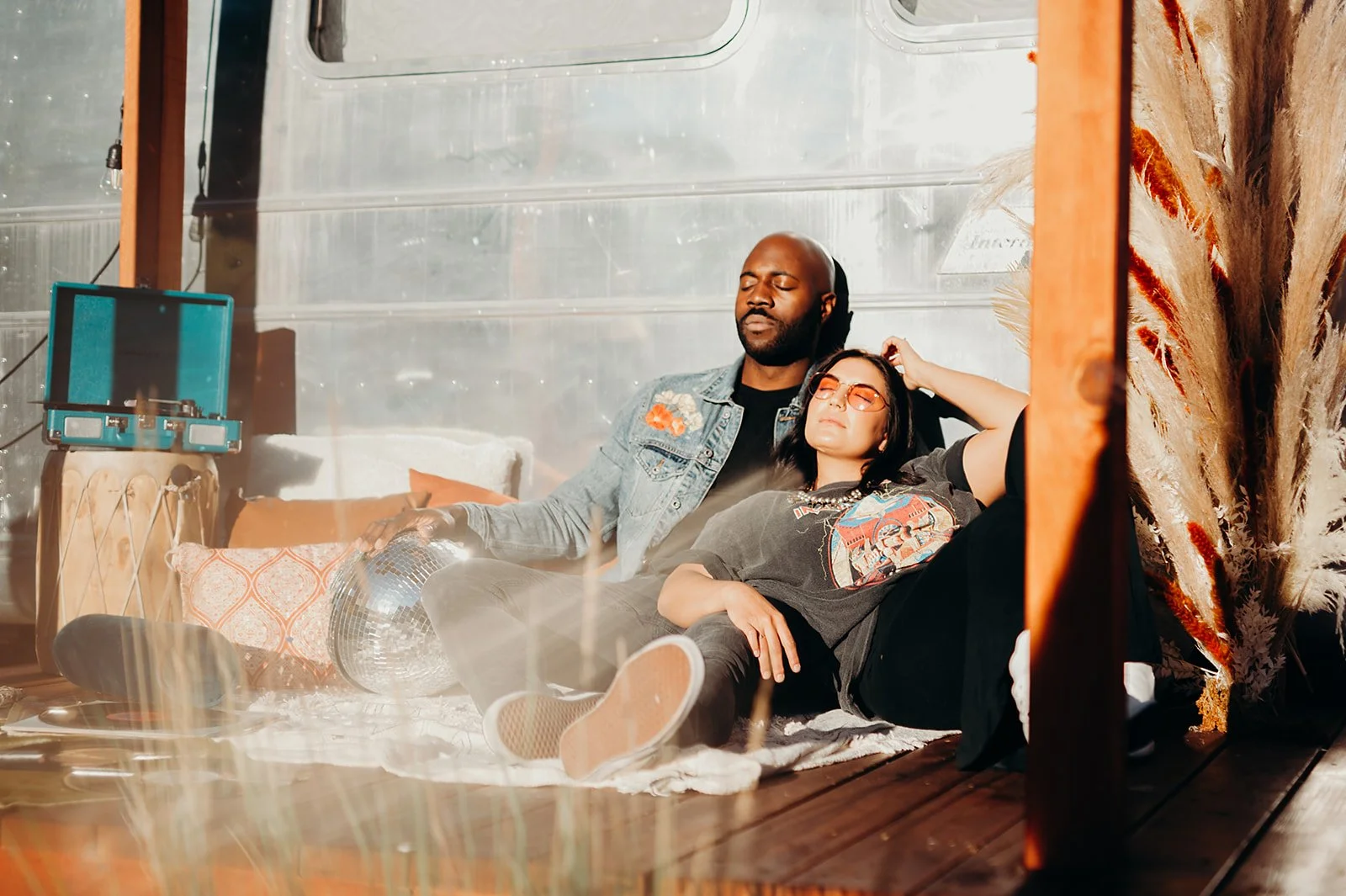 Couple relaxing on a wooden deck with pillows, vintage record player, and disco ball; man in denim jacket, woman in graphic tee and sunglasses, pampas grass decoration nearby.