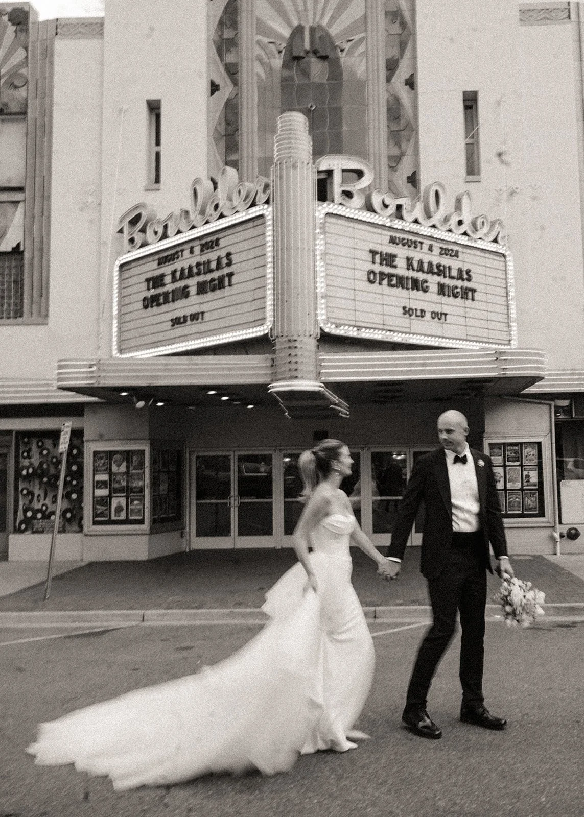 Black and white photo of a couple in wedding attire holding hands in front of a theater with marquee showing "The Kaasilas Opening Night."
