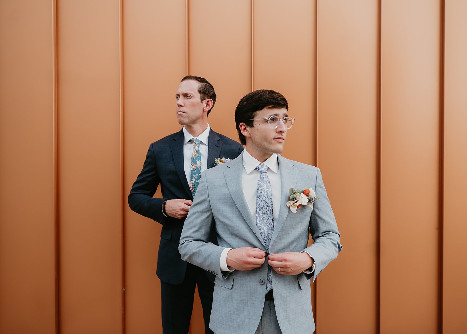 Two men in suits standing in front of an orange panel wall, adjusting their jackets, with floral boutonnieres on their lapels.
