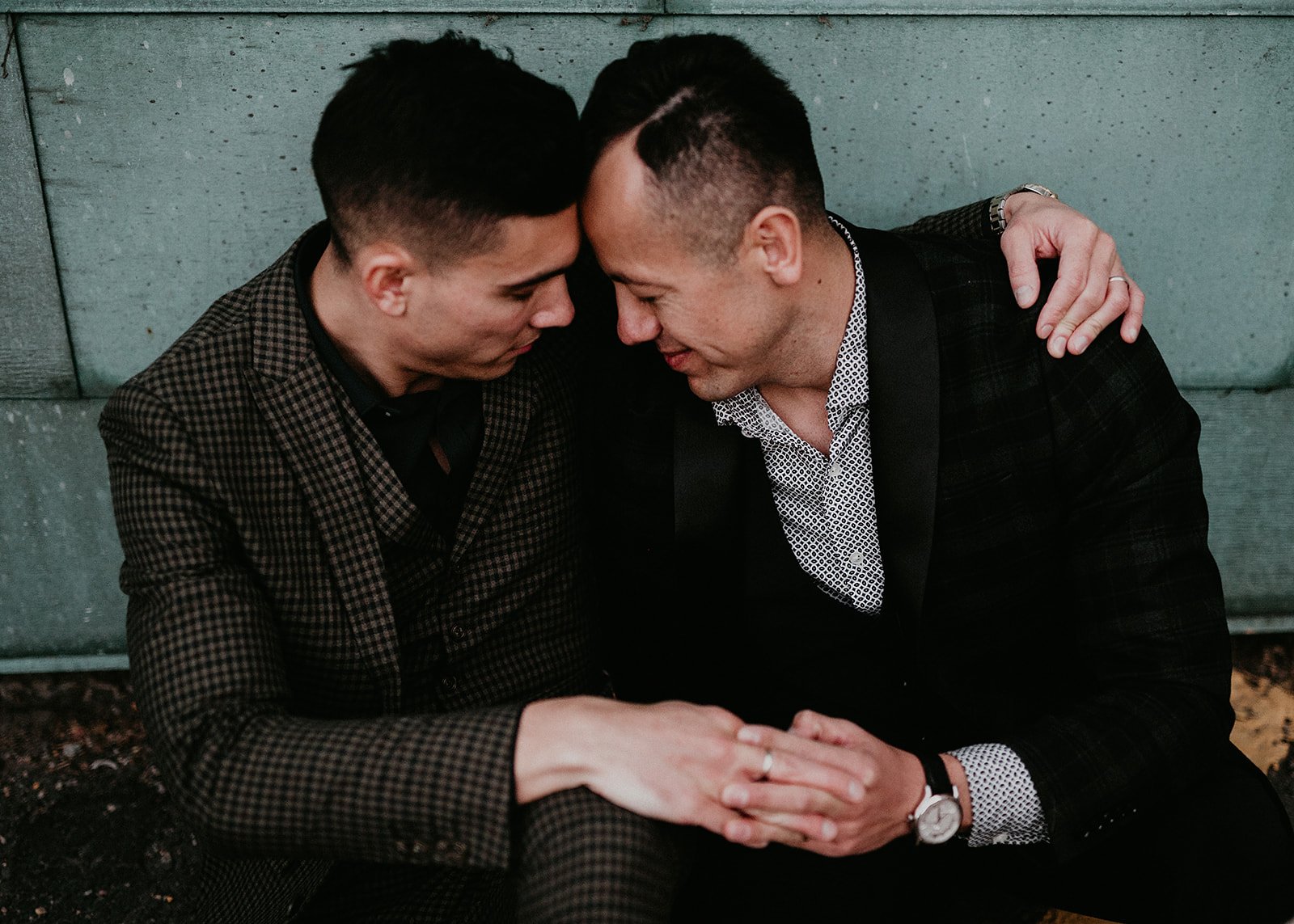 Two people in suits sitting closely, embracing with heads touching, expressing affection.