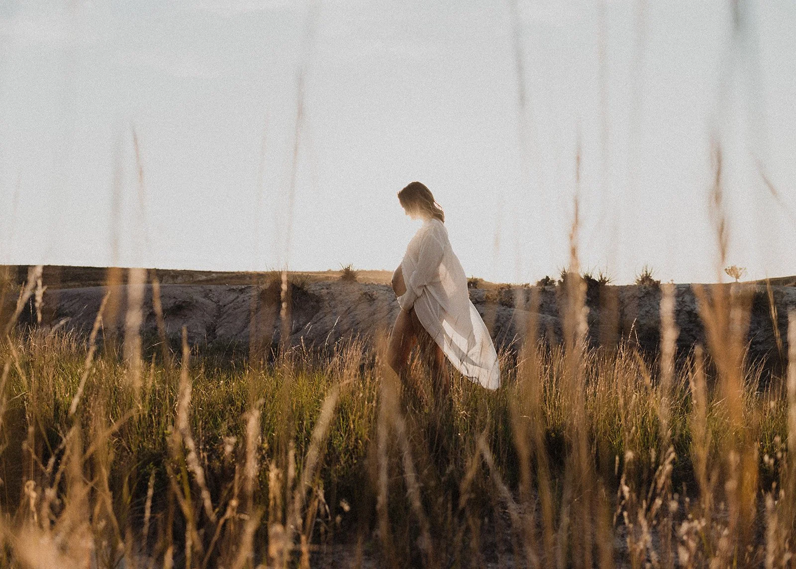 Woman in a light dress walking through tall grass in a field at sunset.