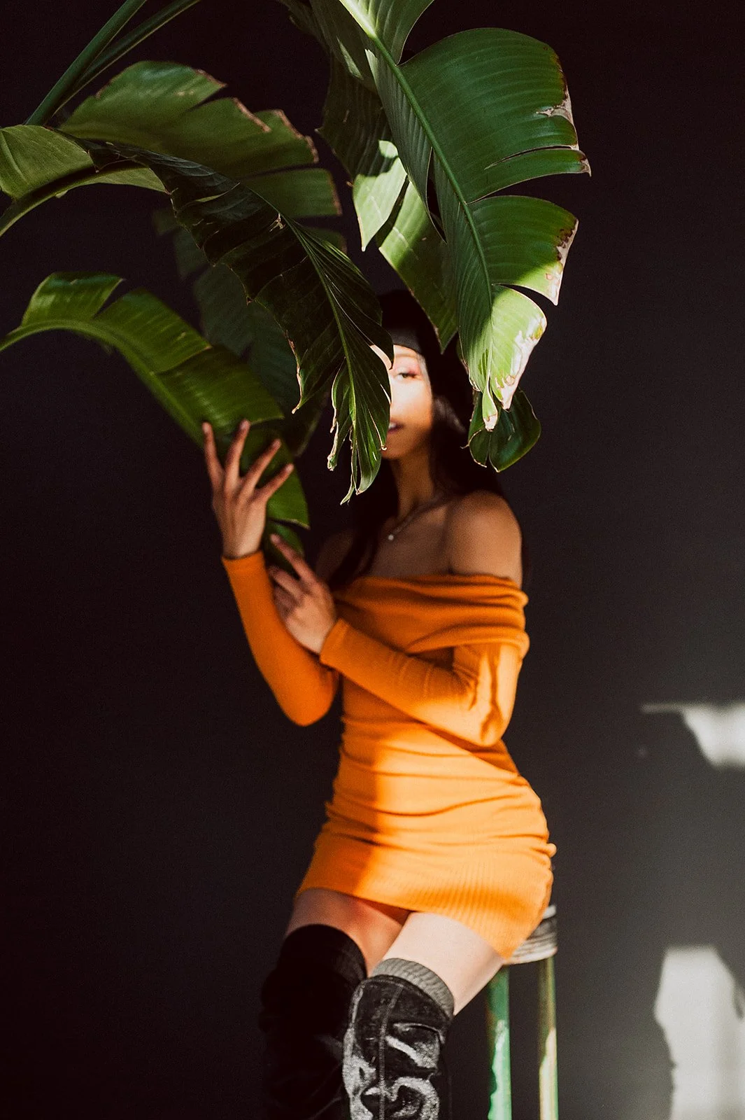 Person in orange dress partially obscured by large green leaves, posing against dark background.