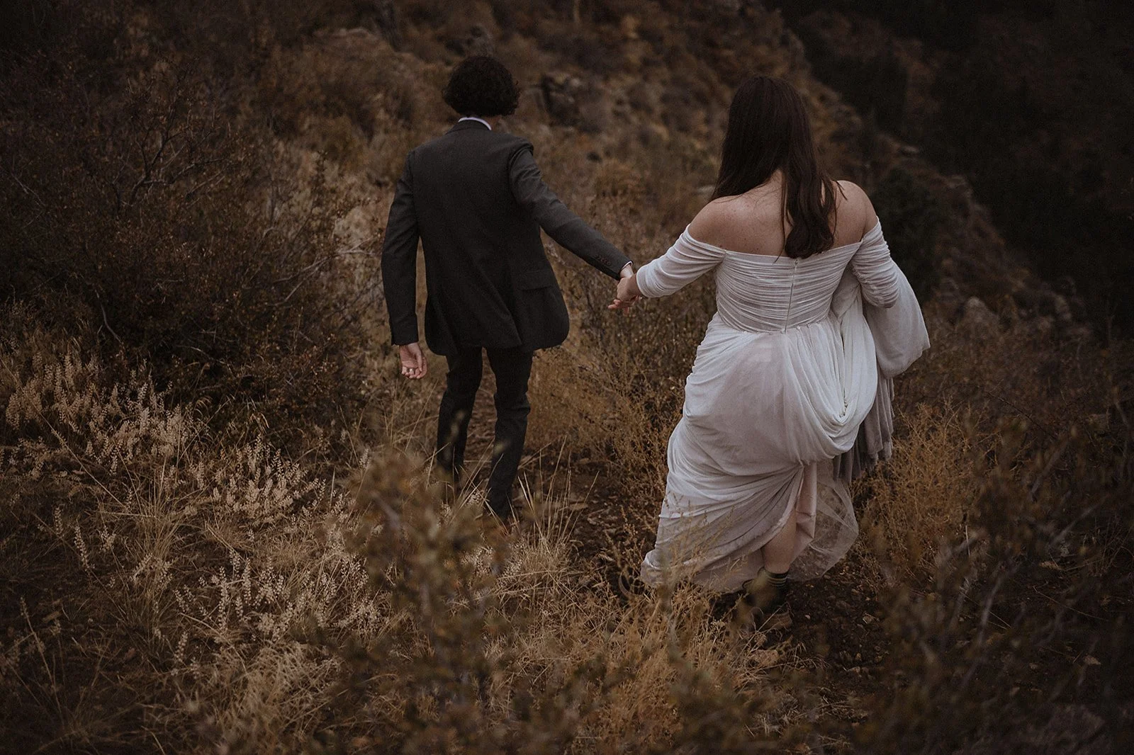 Couple holding hands walking through dry grass on a hillside, dressed in formal attire; woman in a white dress, man in a suit.