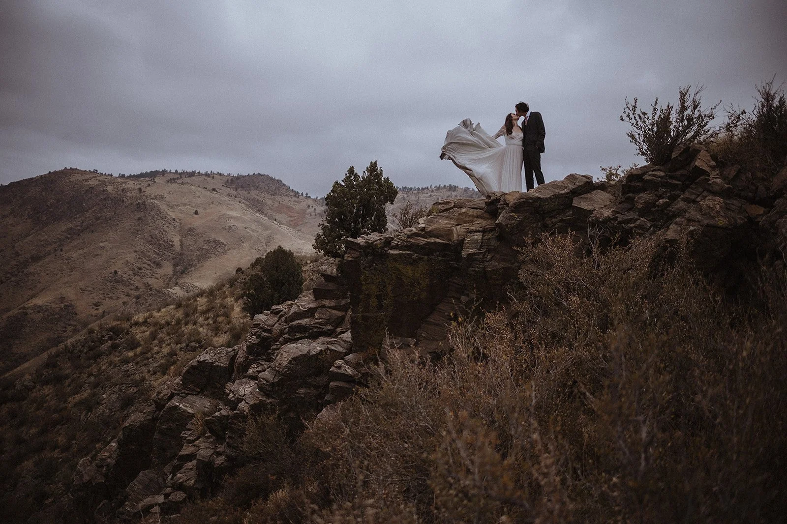Bride and groom standing on a rocky cliff with mountains in the background.