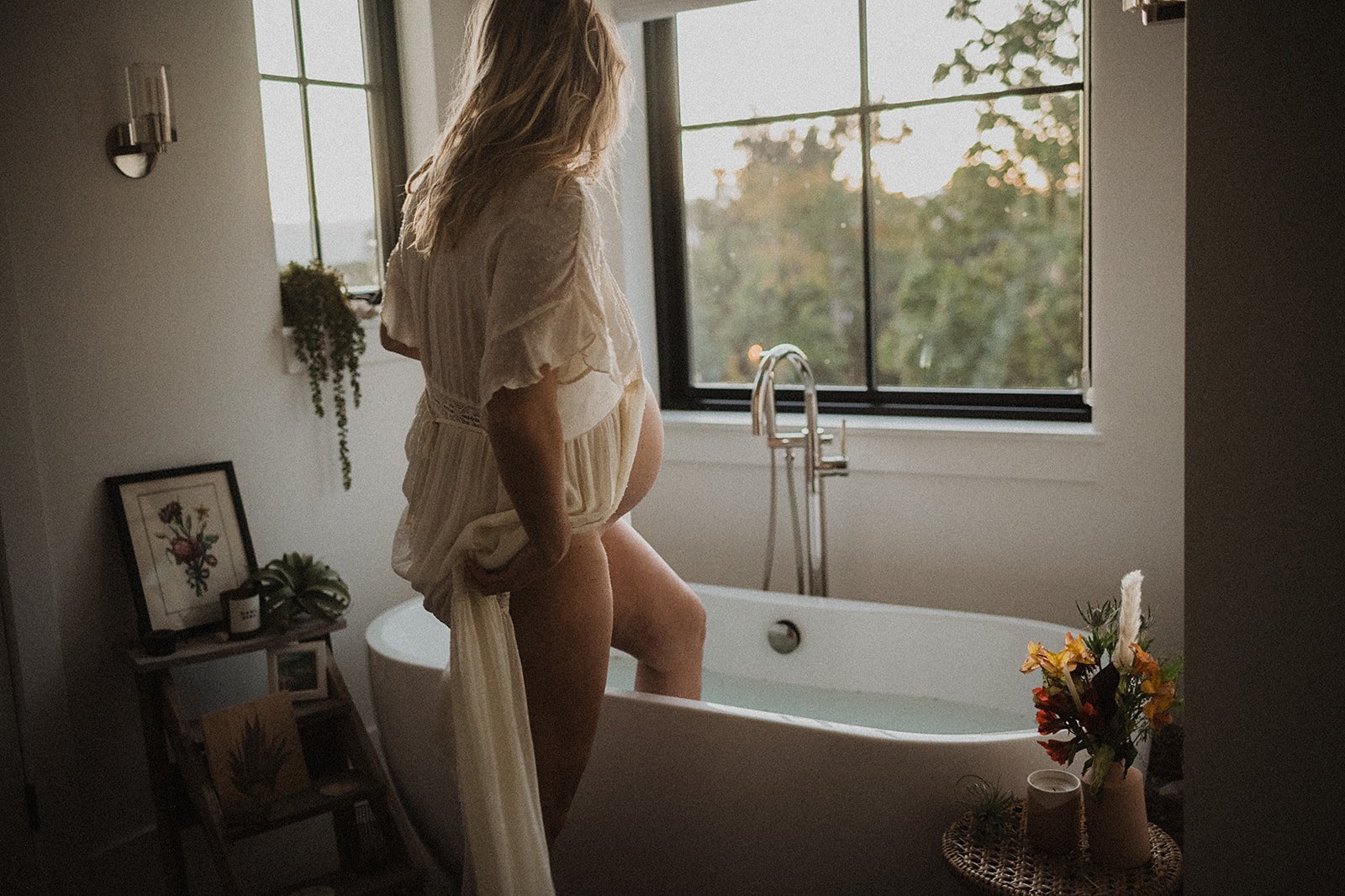 Pregnant woman standing by a bathtub in a bathroom with large windows, surrounded by plants and decor, showcasing a serene setting.