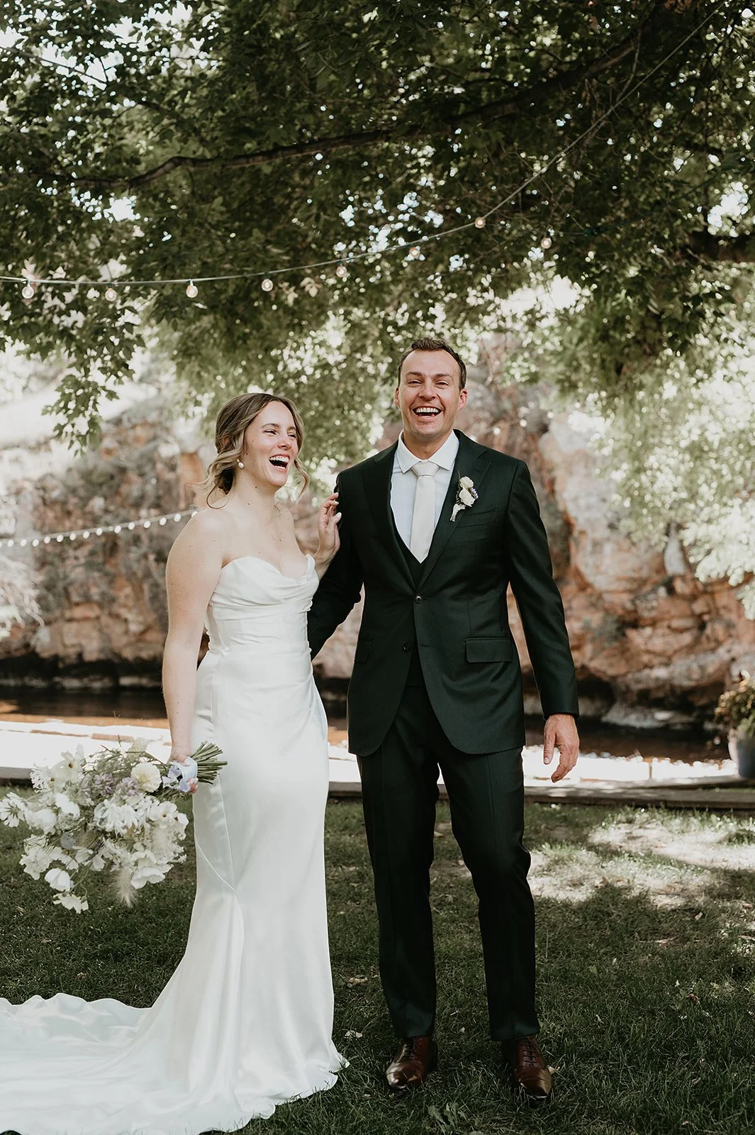 Bride and groom laughing outdoors under tree on wedding day