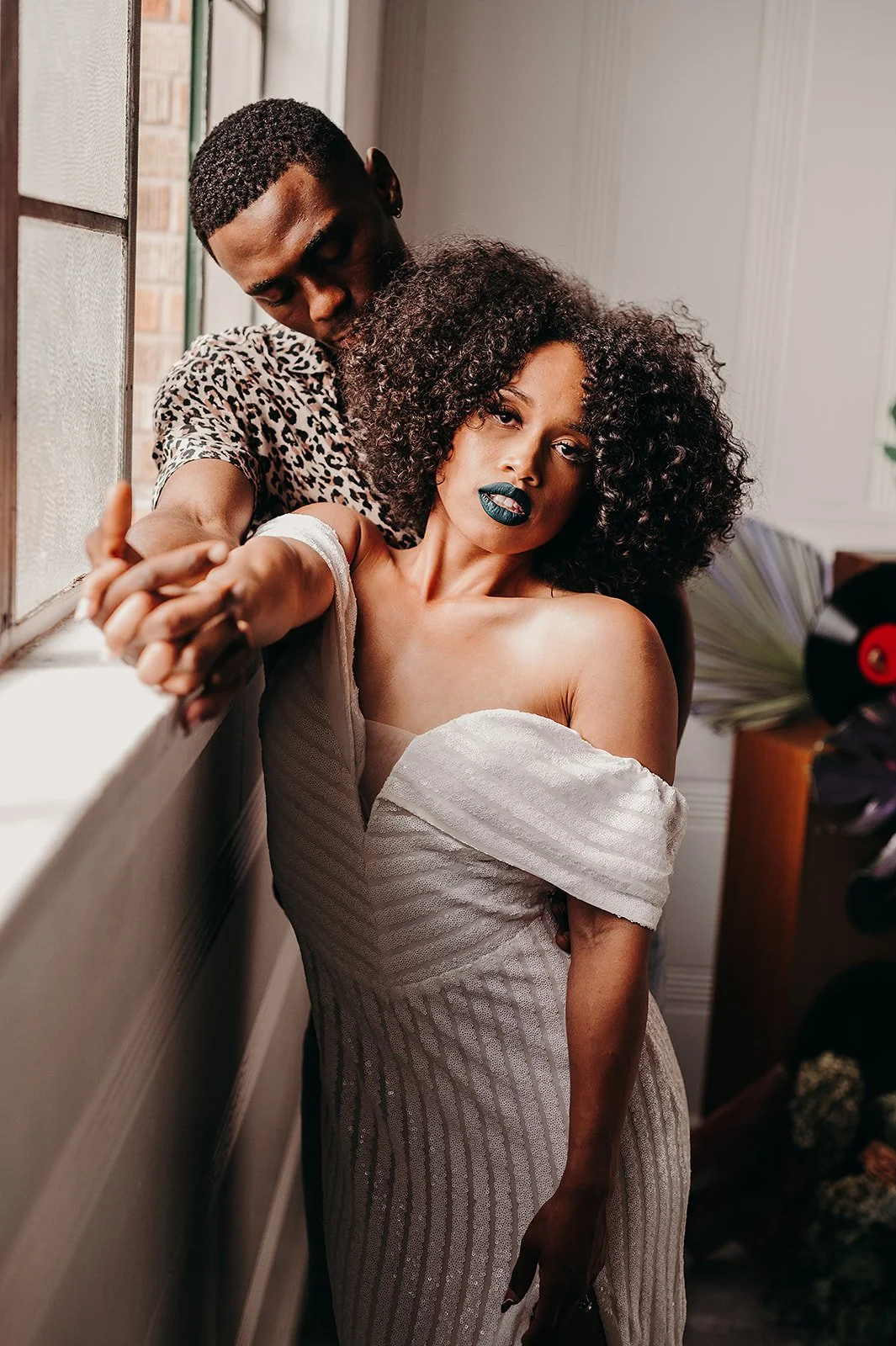A couple in an intimate pose by a window, featuring a woman in a white off-the-shoulder dress with blue lipstick and a man in a leopard print shirt.