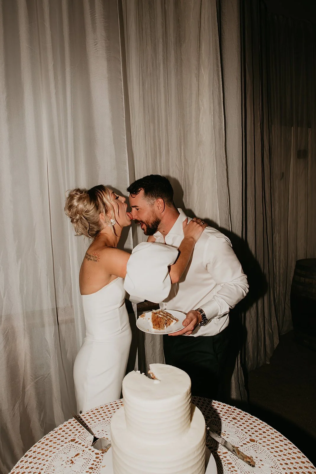 A couple at a wedding reception sharing a piece of cake, with the bride playfully licking the groom's face. A tiered wedding cake is on the table in front of them, covered with a white tablecloth.
