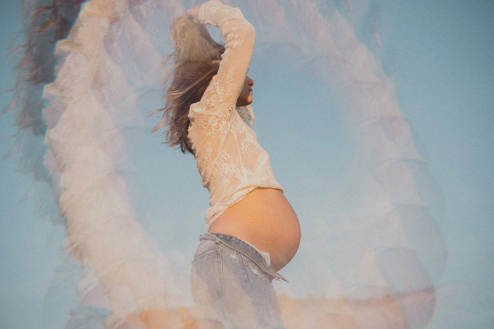 Pregnant woman wearing a lace top and jeans, holding her hair up against a blue sky background, with an artistic translucent overlay effect.