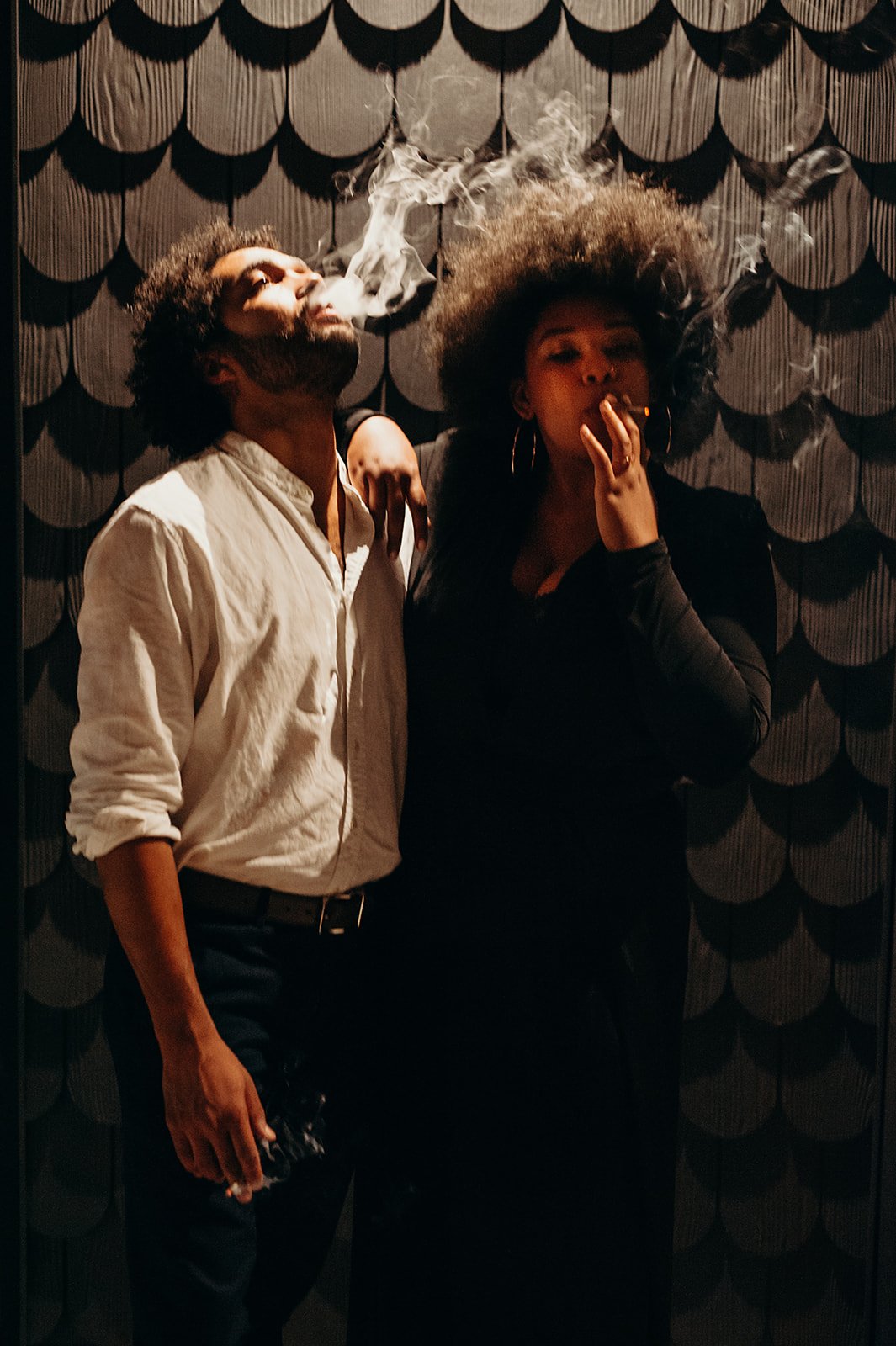Two people exhaling smoke against a textured dark background, one wearing a white shirt and the other in black clothing.