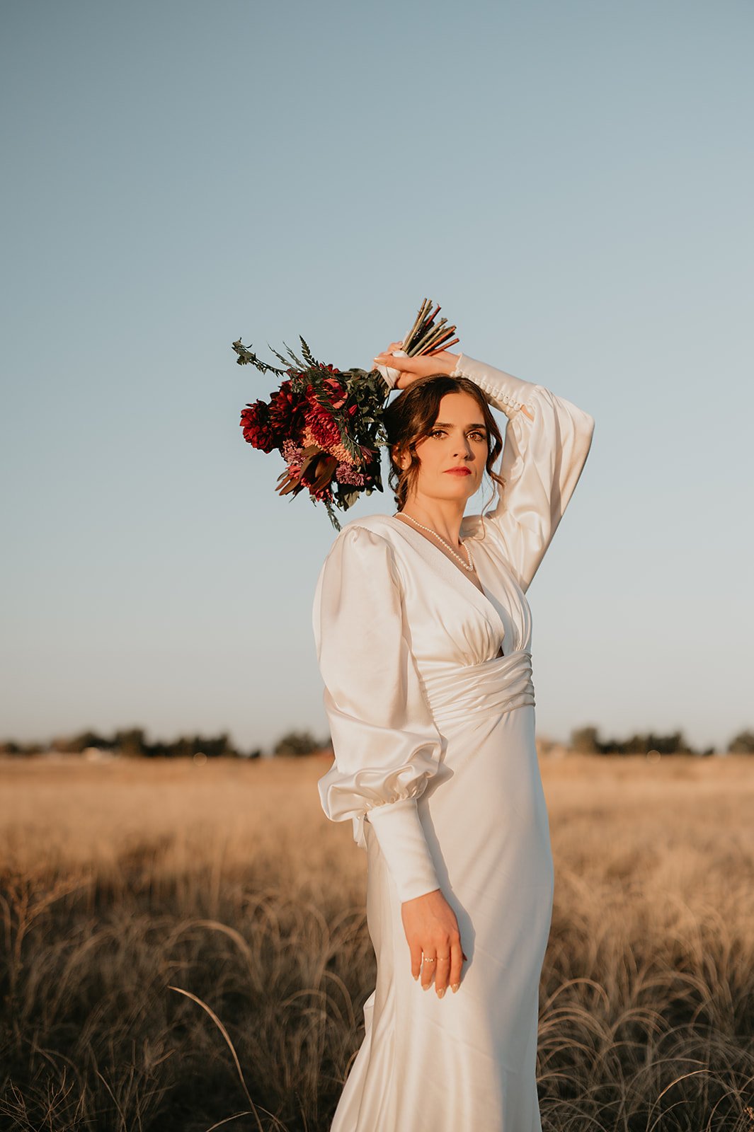 Bride in a satin wedding dress with puffed sleeves holding a bouquet of burgundy flowers against a field background.