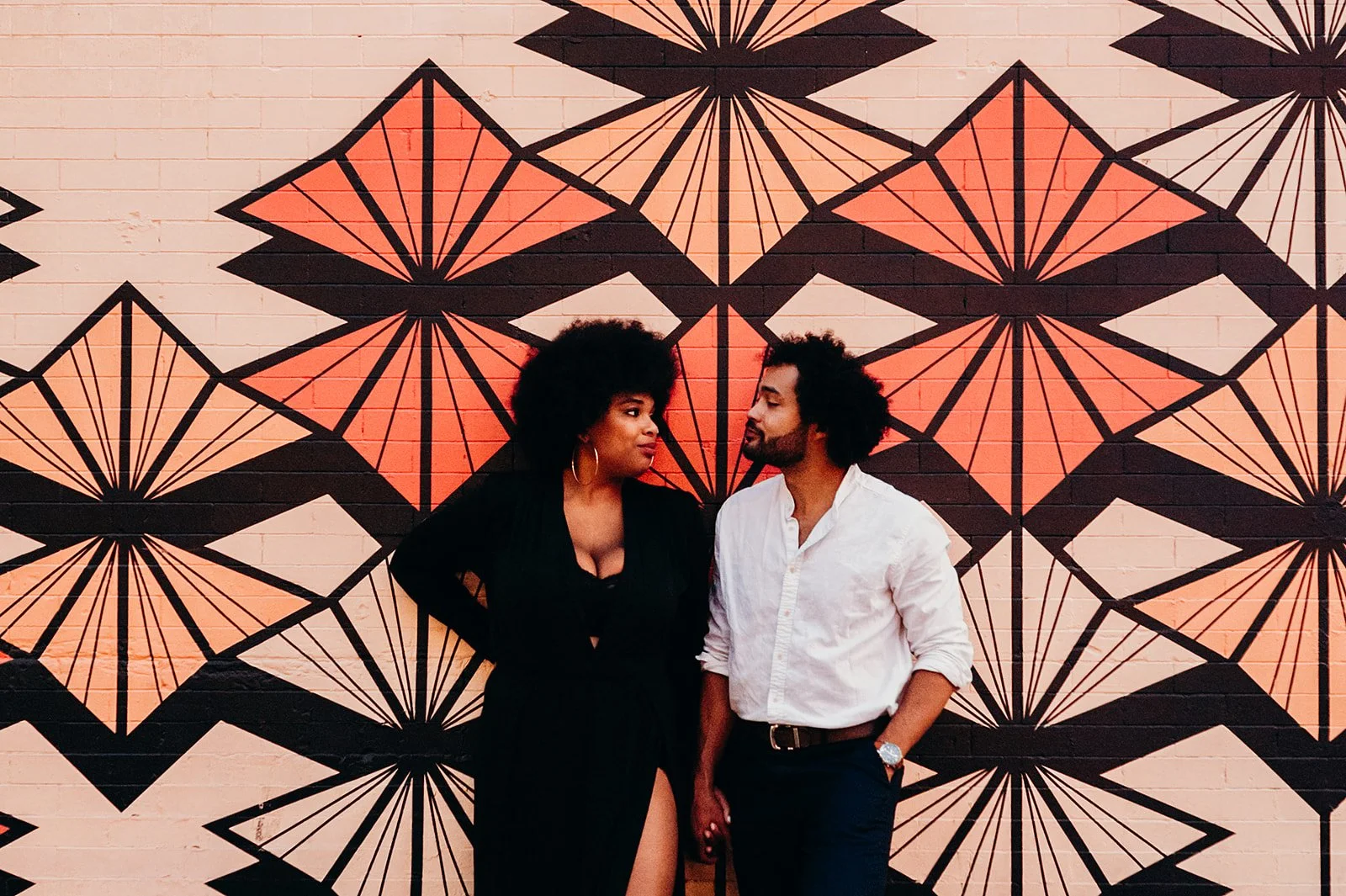 A couple standing in front of a geometric patterned mural. The wall features a design with black, red, and orange shapes. The woman is wearing a black dress, and the man is in a white shirt and dark pants, looking at each other.