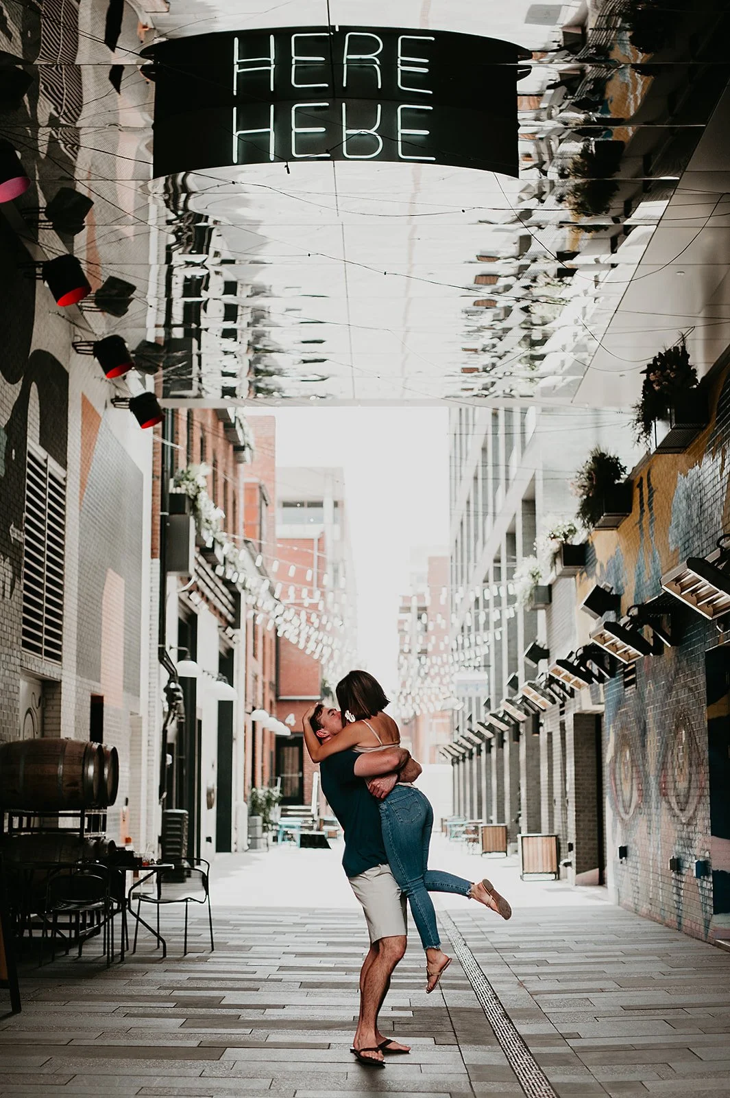 Couple embracing under "HERE" sign in urban alley with string lights.