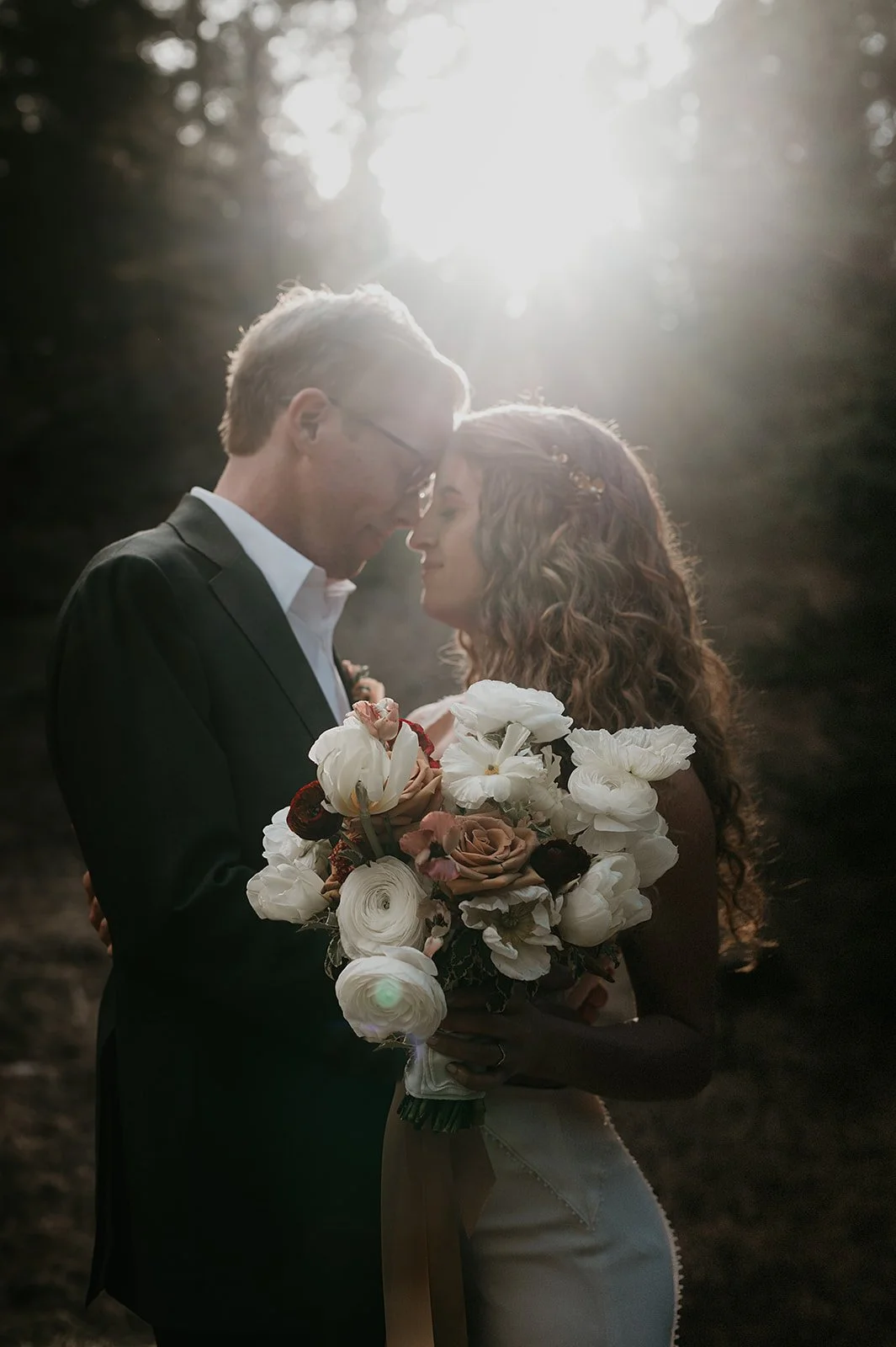 A couple in formal attire sharing an intimate moment in a sunlit outdoor setting, with the woman holding a bouquet of white and pink flowers.