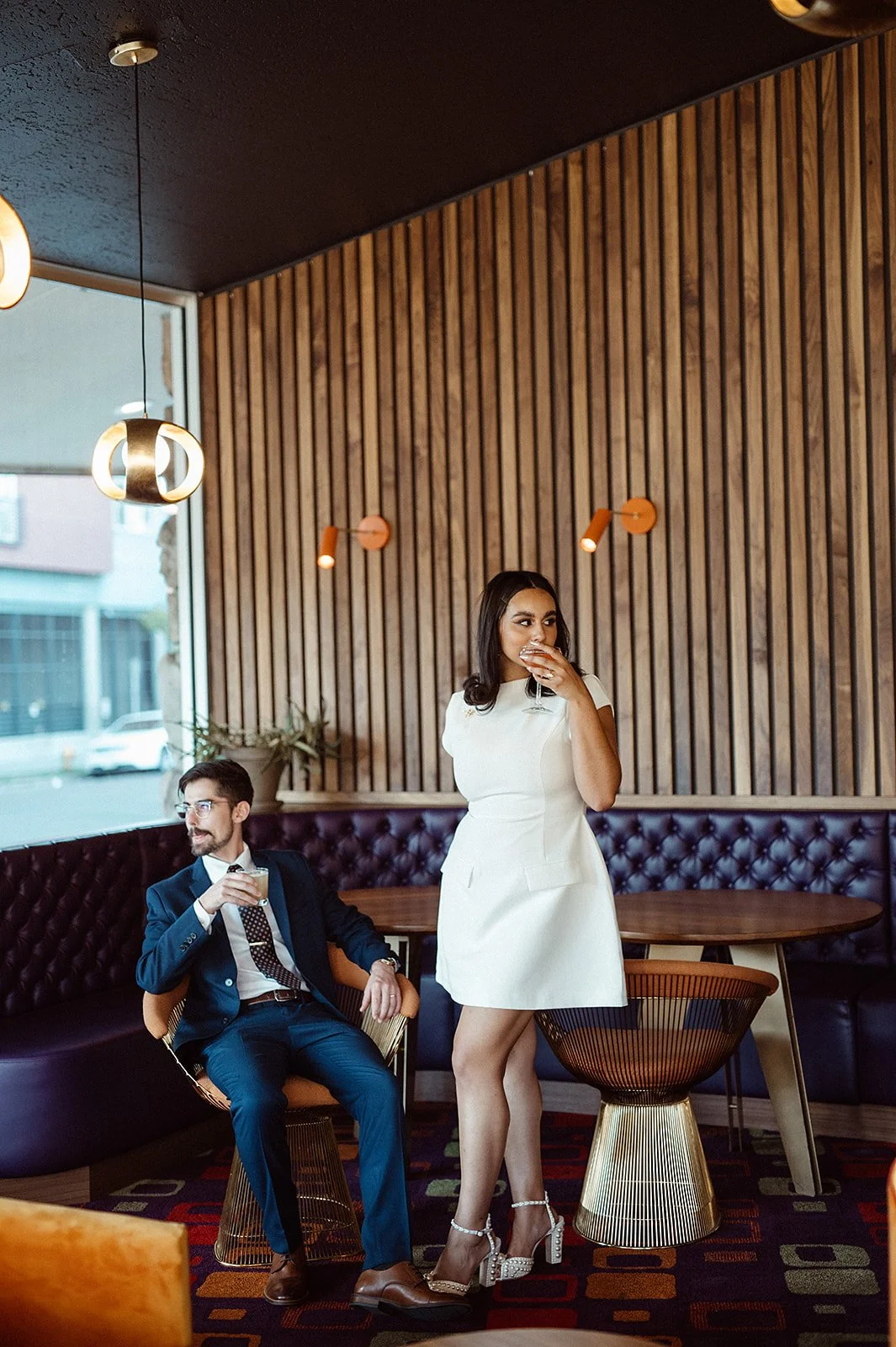 A man in a suit sitting and a woman in a white dress standing in a stylish modern lounge, both holding drinks. The room features wooden panel walls and unique lighting fixtures.