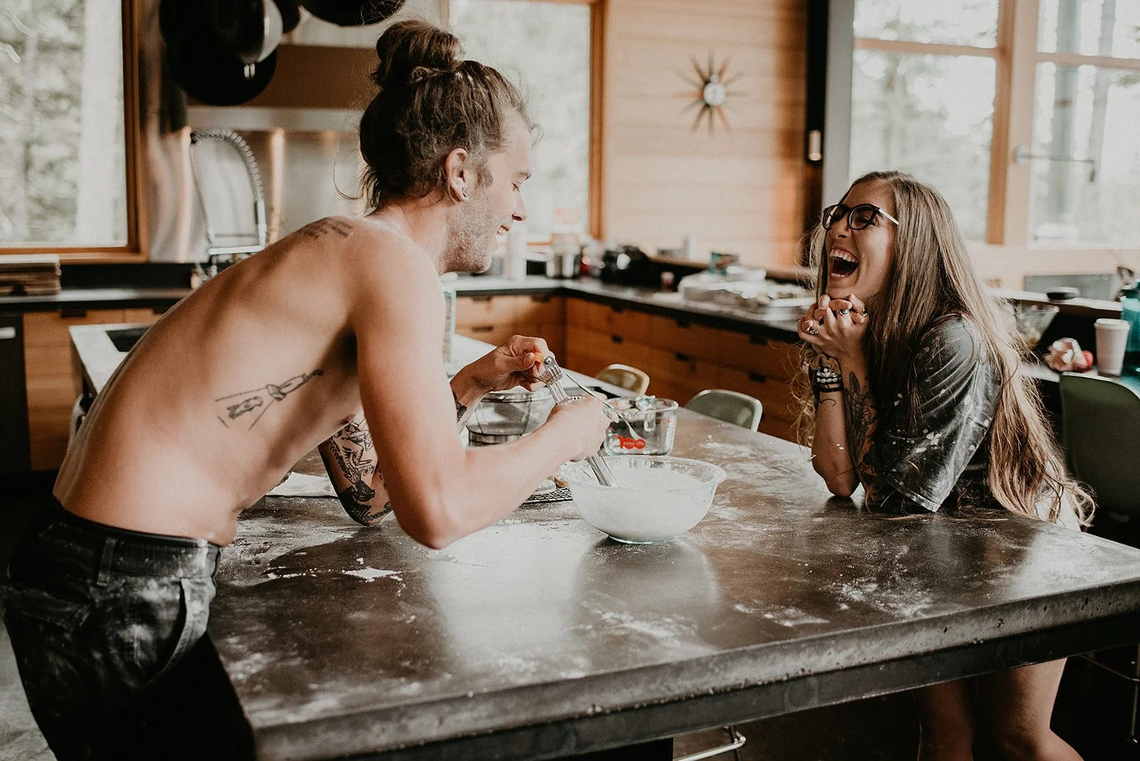 Two people laughing and baking together in a kitchen, with one person shirtless, mixing batter in a bowl on a countertop.