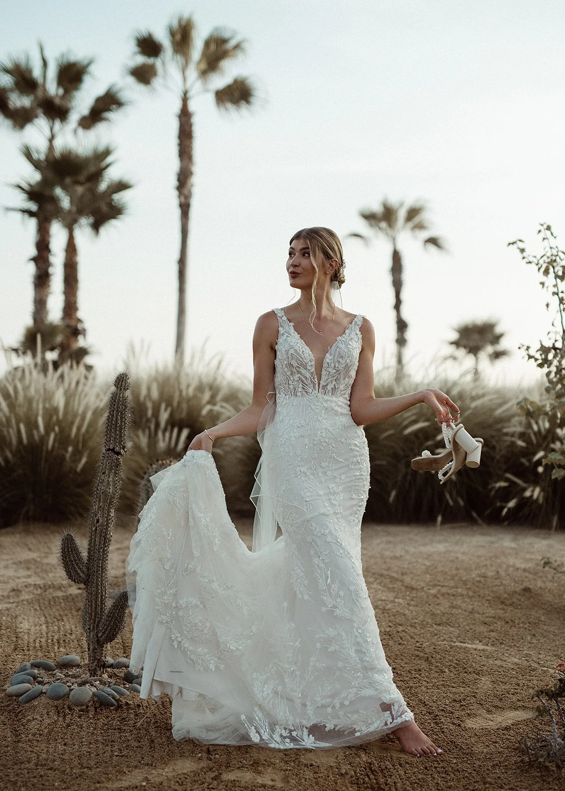Bride in a white wedding dress holding shoes, standing in a desert landscape with palm trees and cactus.