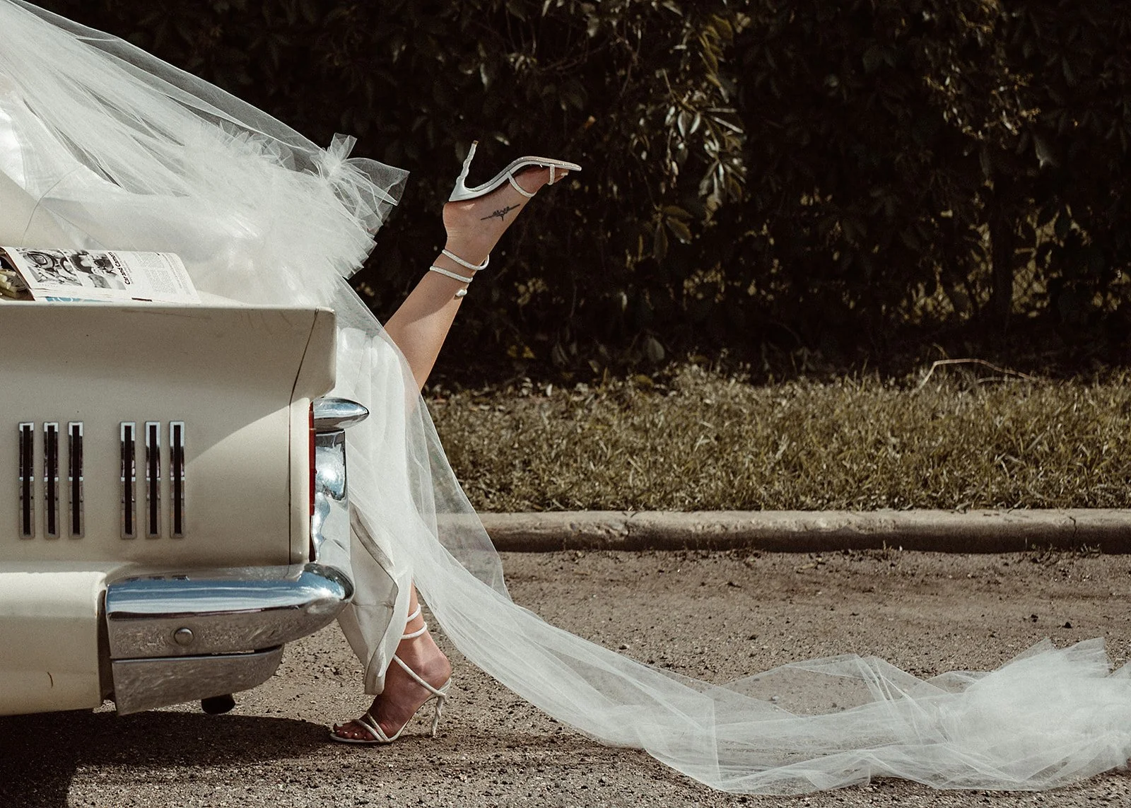 Bride in white dress and heels with leg sticking out of a vintage car.